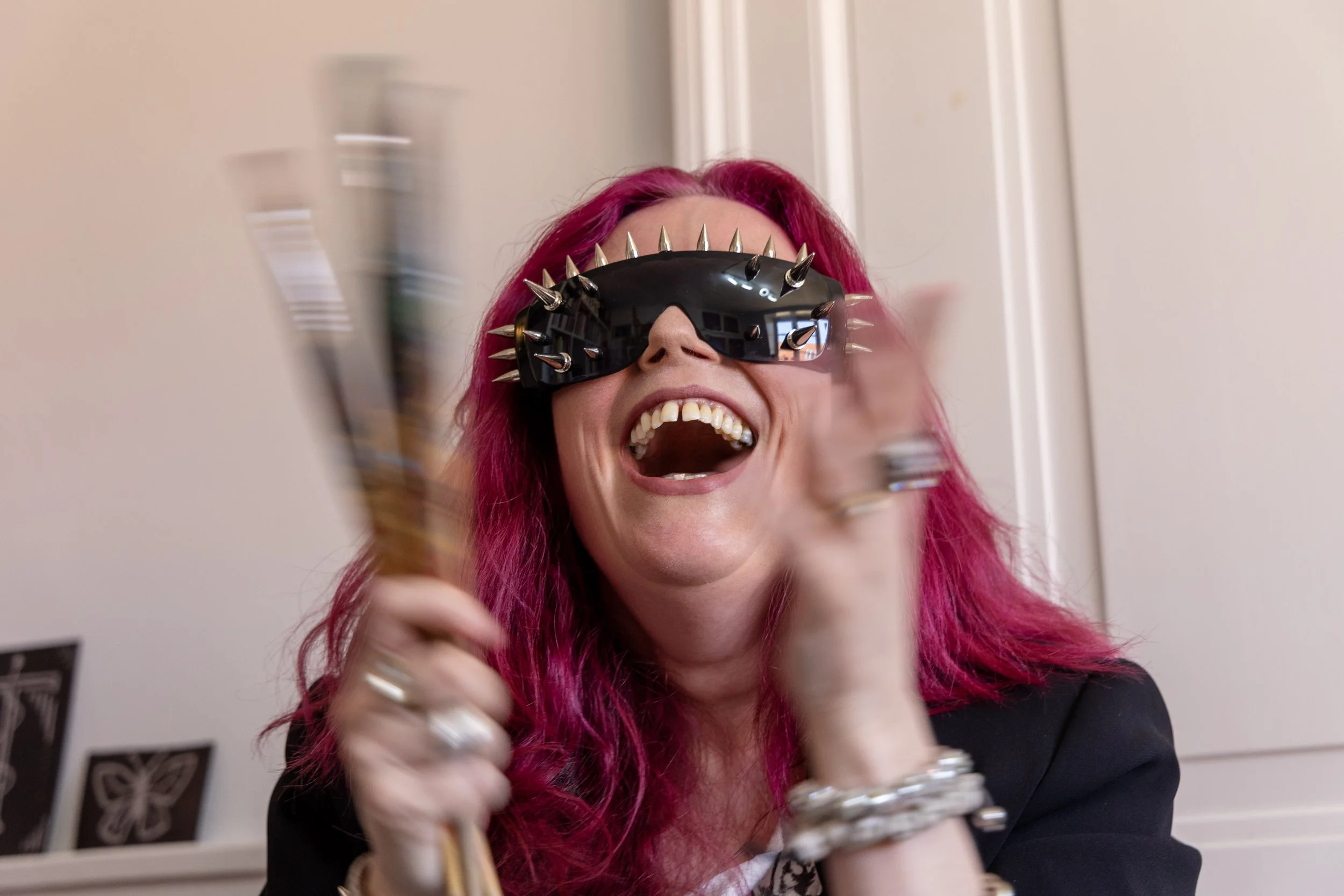 A woman with bright pink hair laughing, wearing black futuristic sunglasses with silver spikes, holding a paintbrush, in a room with white walls and butterfly artwork.