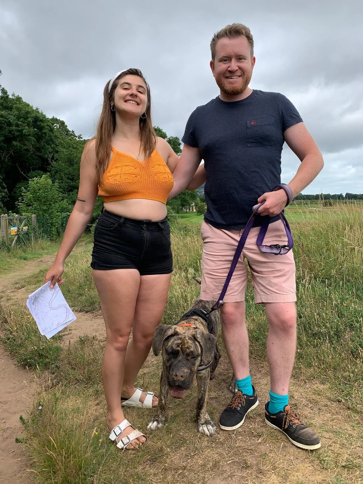A young woman and a young man standing on a dirt path outdoors during daytime, smiling at the camera, with a brindle-coated dog on a leash. The woman is wearing a yellow crop top, black shorts, and white sandals. The man is wearing a navy T-shirt, light pink shorts, and dark sneakers with bright blue socks. She is holding a map in her left hand, and his arm is around her shoulder. The background consists of green trees and grasses under a cloudy sky.