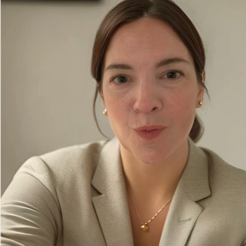 A woman with dark hair tied back, wearing a beige blazer and gold earrings, looks directly at the camera. She appears thoughtful and focused.