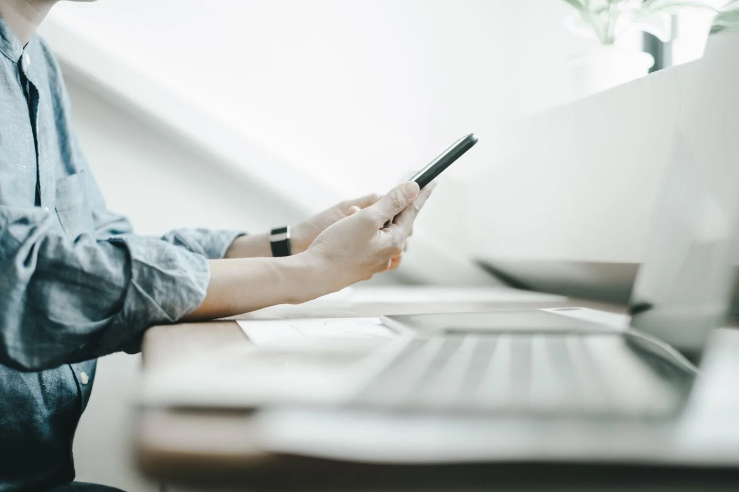 person seated at office desk with papers and a computer on it, looking at smart phone