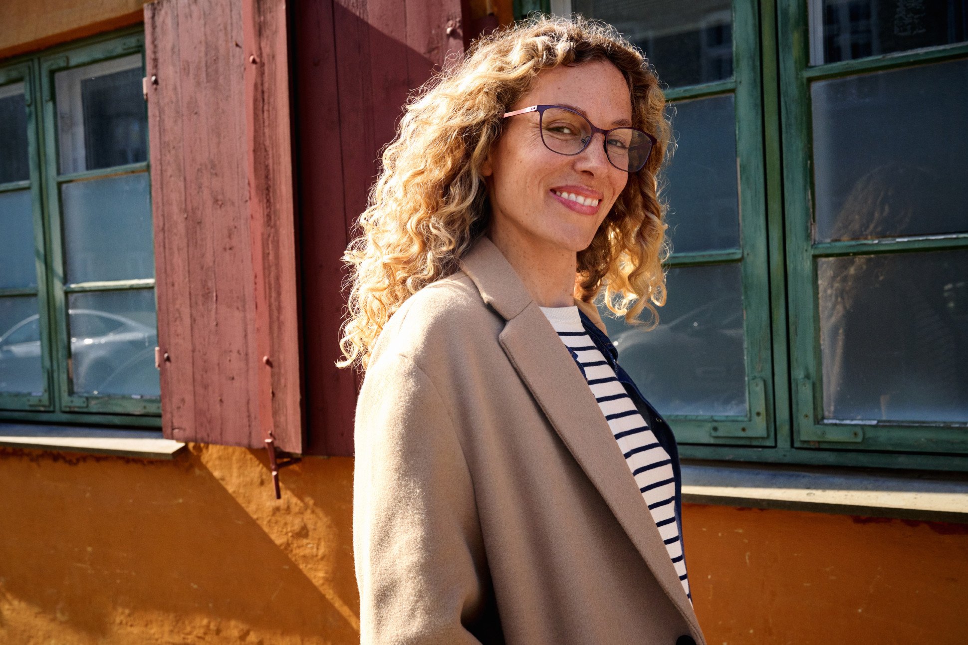 A woman with curly blonde hair wearing glasses, a beige blazer, and a striped shirt standing outdoors in front of a vibrant, weathered building with green window frames and a red-brown exterior.
