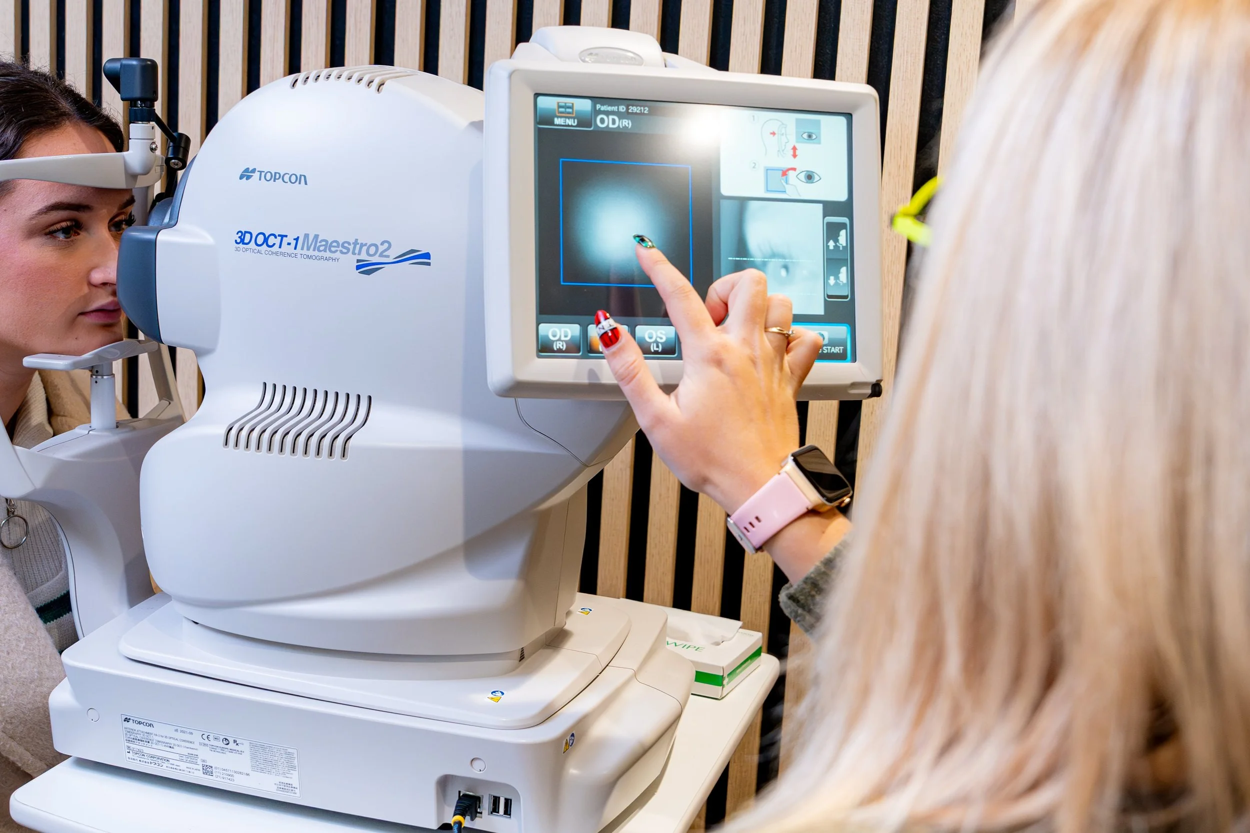 A person is operating a large medical imaging machine, likely an eye scanner, while another woman is positioned to undergo an eye exam.