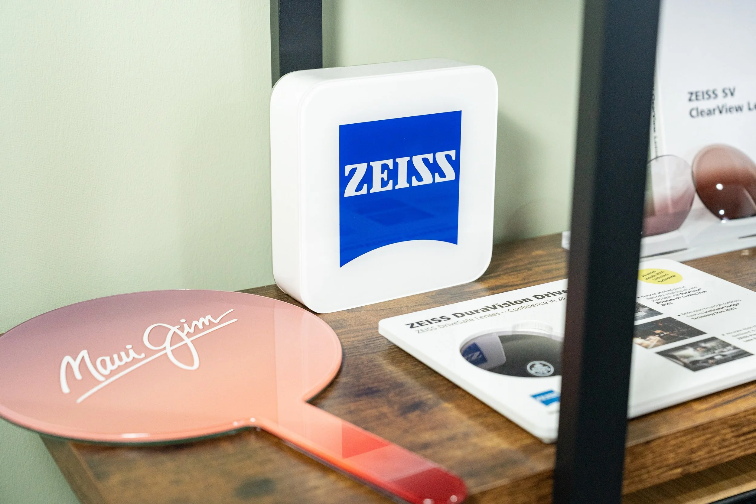 Display of Zeiss branding materials, including a box with Zeiss logo, a pink paddle with 'Maui Jim' text, and a brochure about Zeiss DriveLens on a wooden surface.