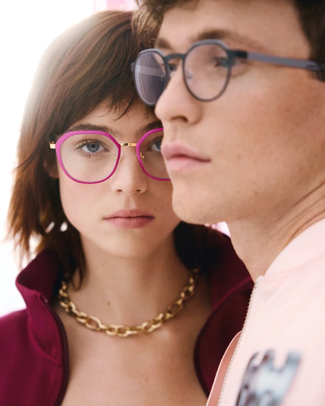 A close-up of a young woman with short brown hair and wearing pink eyeglasses, and a young man with glasses. The woman is looking directly at the camera while the man faces sideways.
