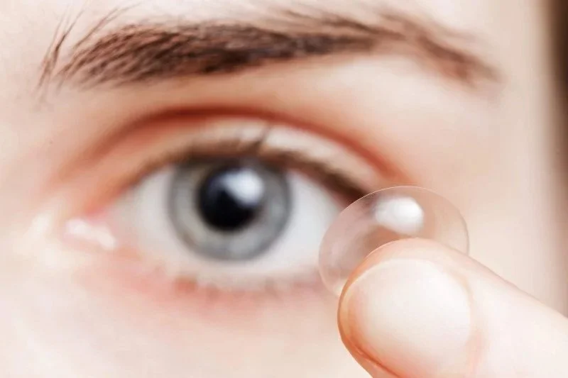 A close-up of an eye with a hand holding a contact lens near it, ready to be placed on the eye.