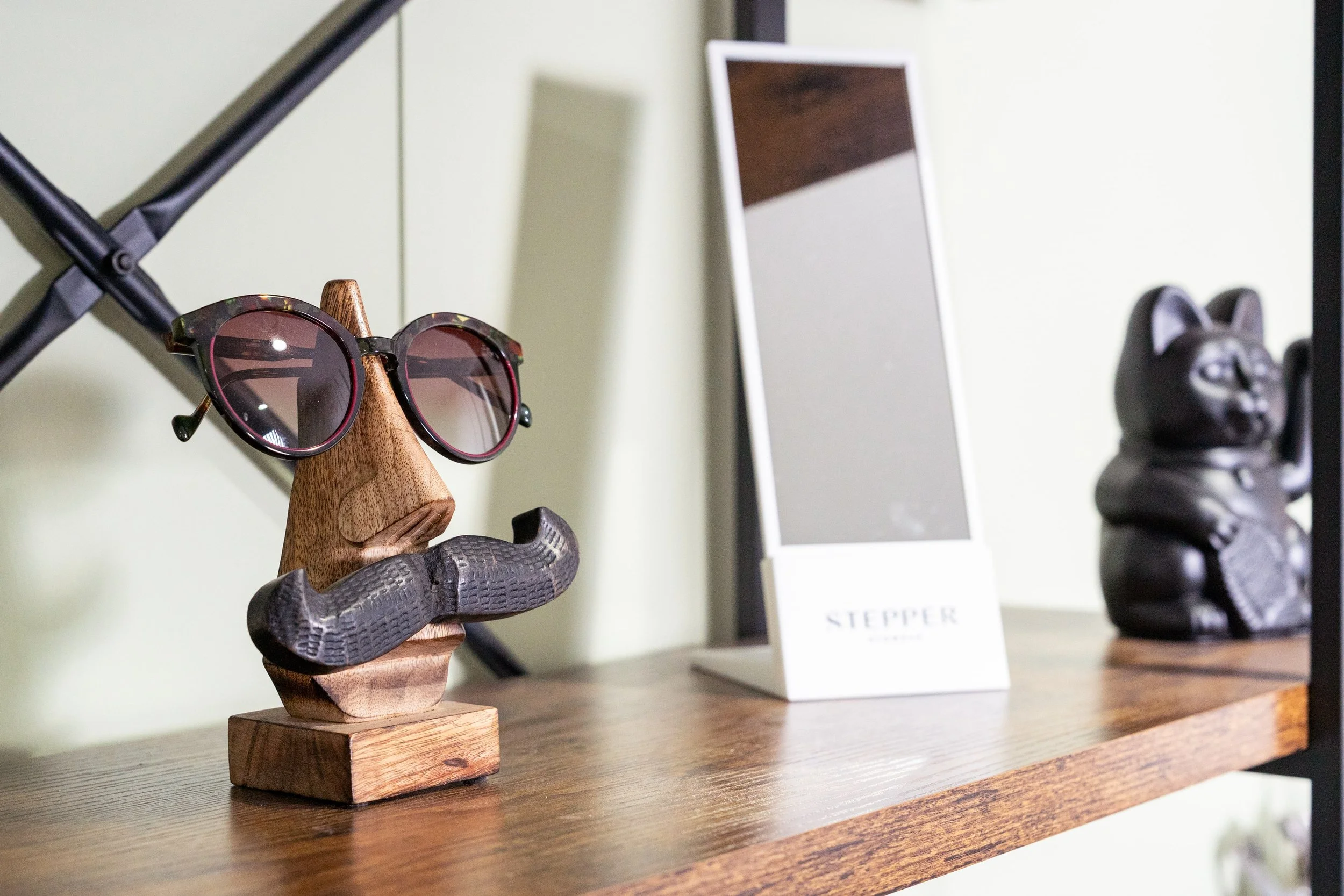 Decorative glasses stand with sunglasses on a wooden nose, framed mirror, and a black lucky cat figurine on a wooden shelf.