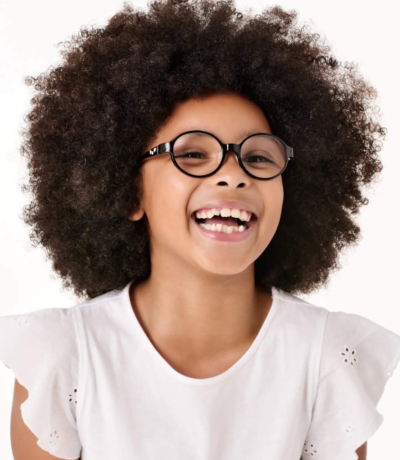 A young girl with curly hair wearing glasses and a white shirt, smiling broadly showing missing teeth.