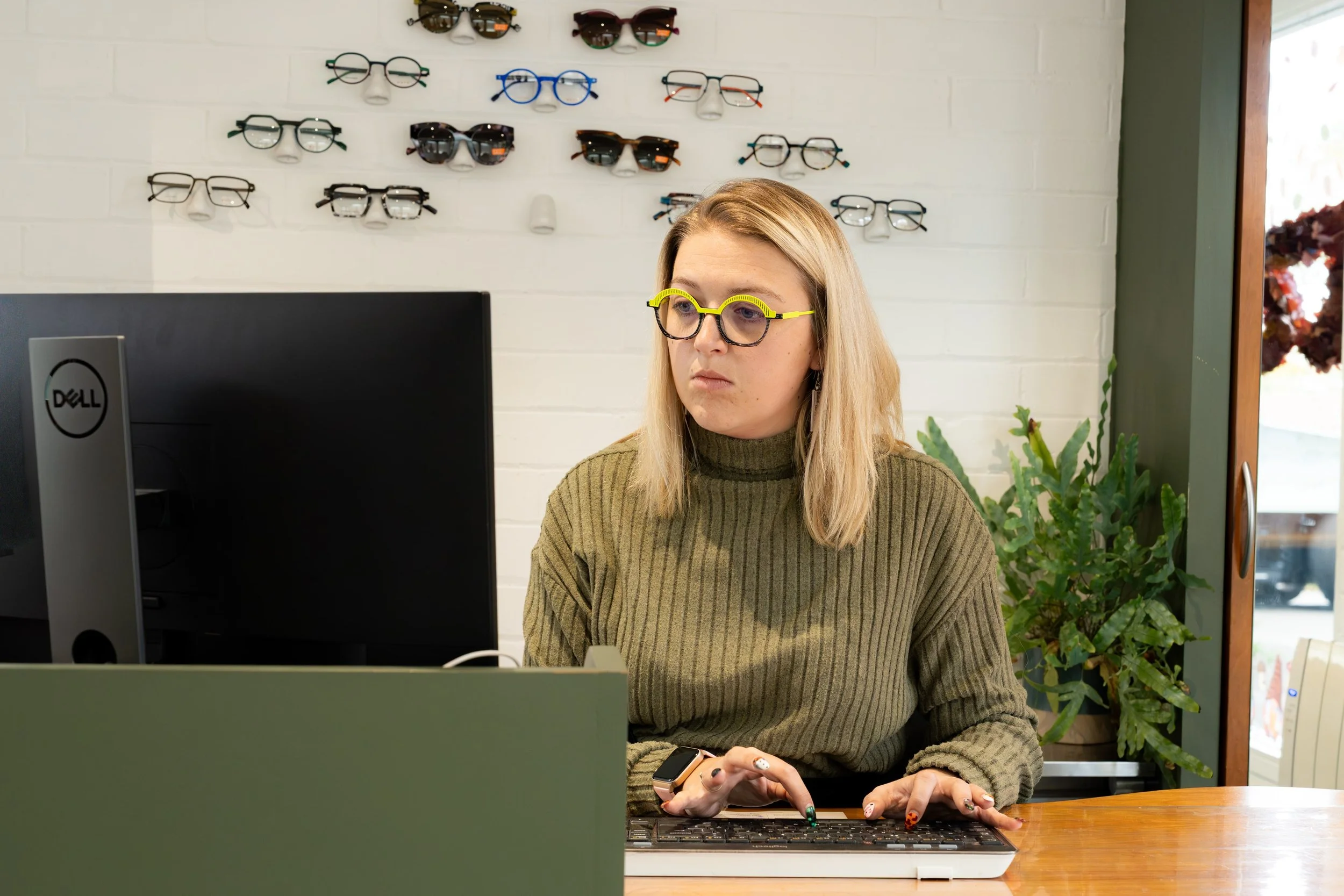 A woman sitting at a desk working on a computer in an optometry office, with multiple eyeglasses displayed on the white brick wall behind her.