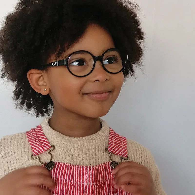 Young child with curly hair and glasses smiles, wearing a cream sweater and red-striped overalls against a plain light background.