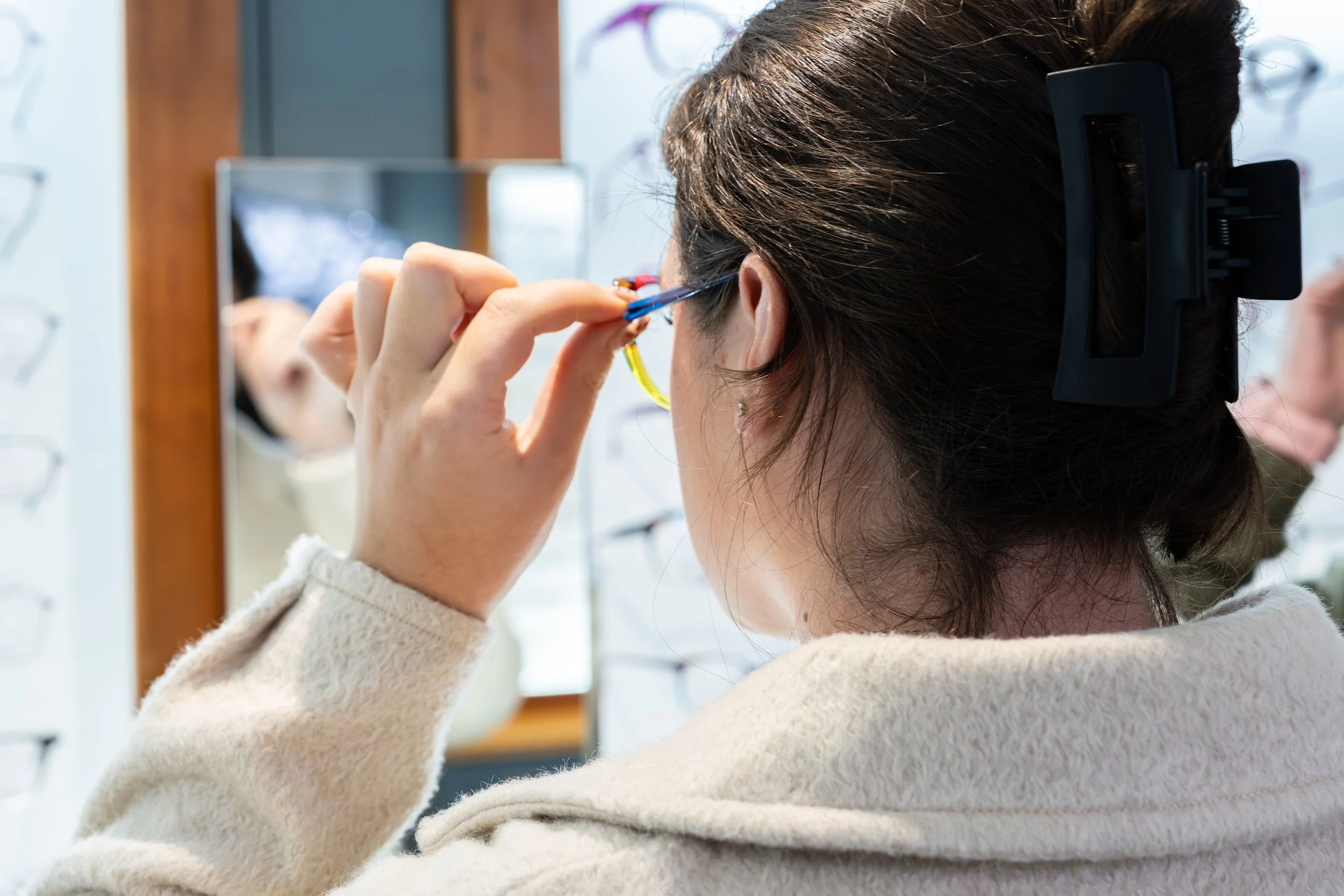 A woman with dark hair secured by a black hair clip is trying on a pair of glasses at an optical shop, reflected in a mirror in front of her.