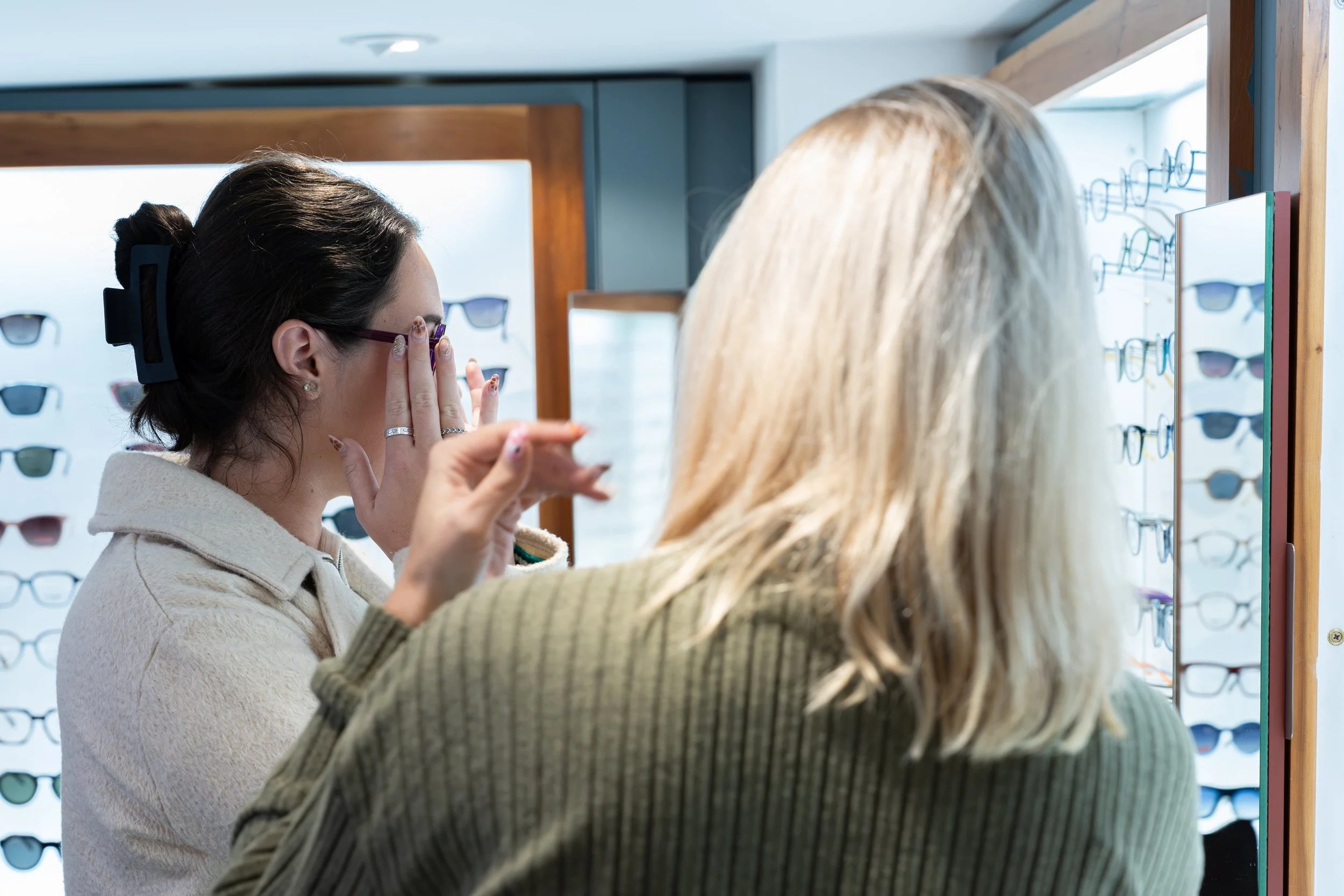 Two women shopping for sunglasses at an eyewear store, with sunglass displays in the background.