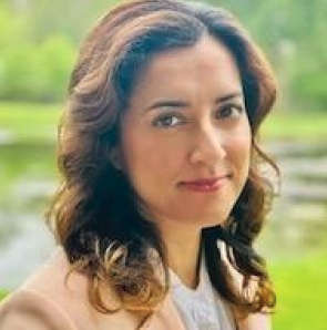 A woman with brown wavy hair outdoors near a body of water, wearing a tan blazer and white top.