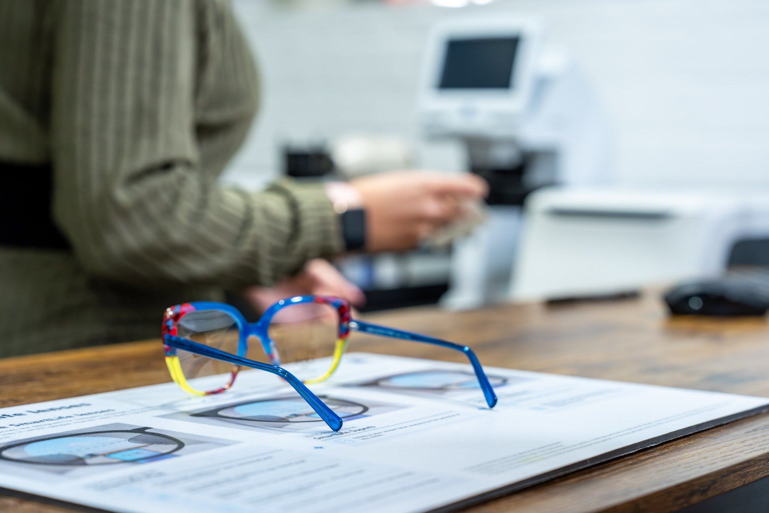 Colorful children's eyeglasses resting on a newspaper on a wooden desk, with a person in the background using a tablet in an office setting.
