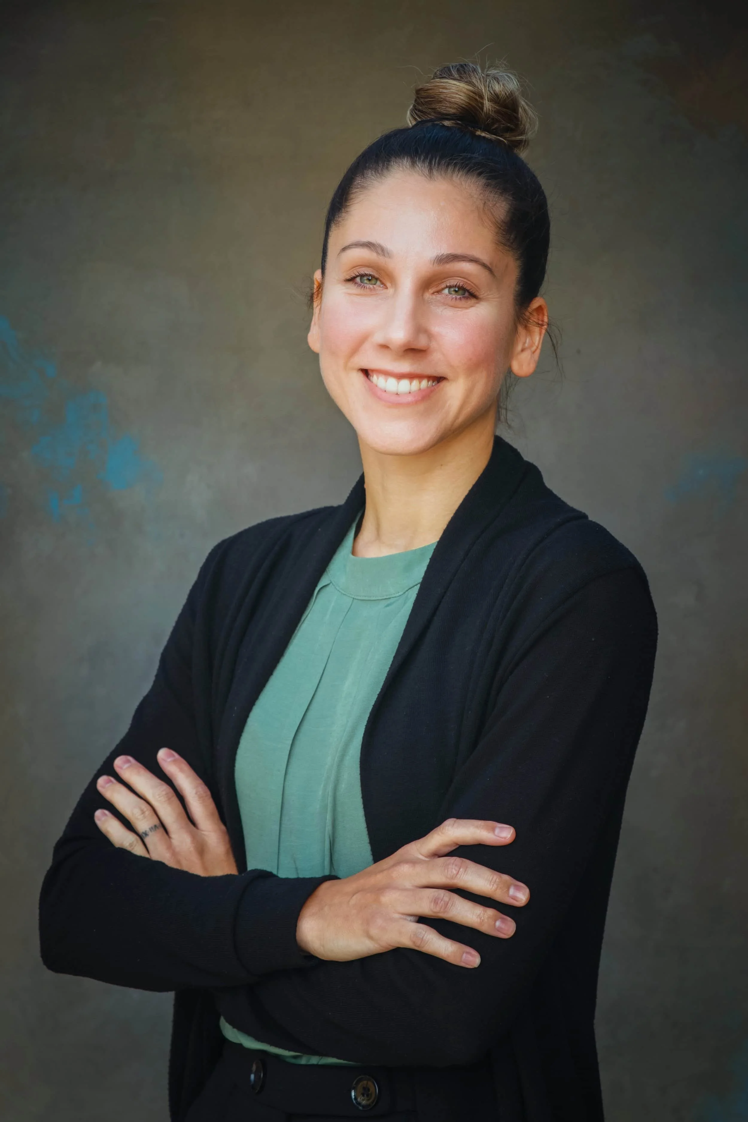 Jessica Cardona, functional practitioner, smiles with arms crossed, hair in a bun, in green top and black cardigan against a neutral background.