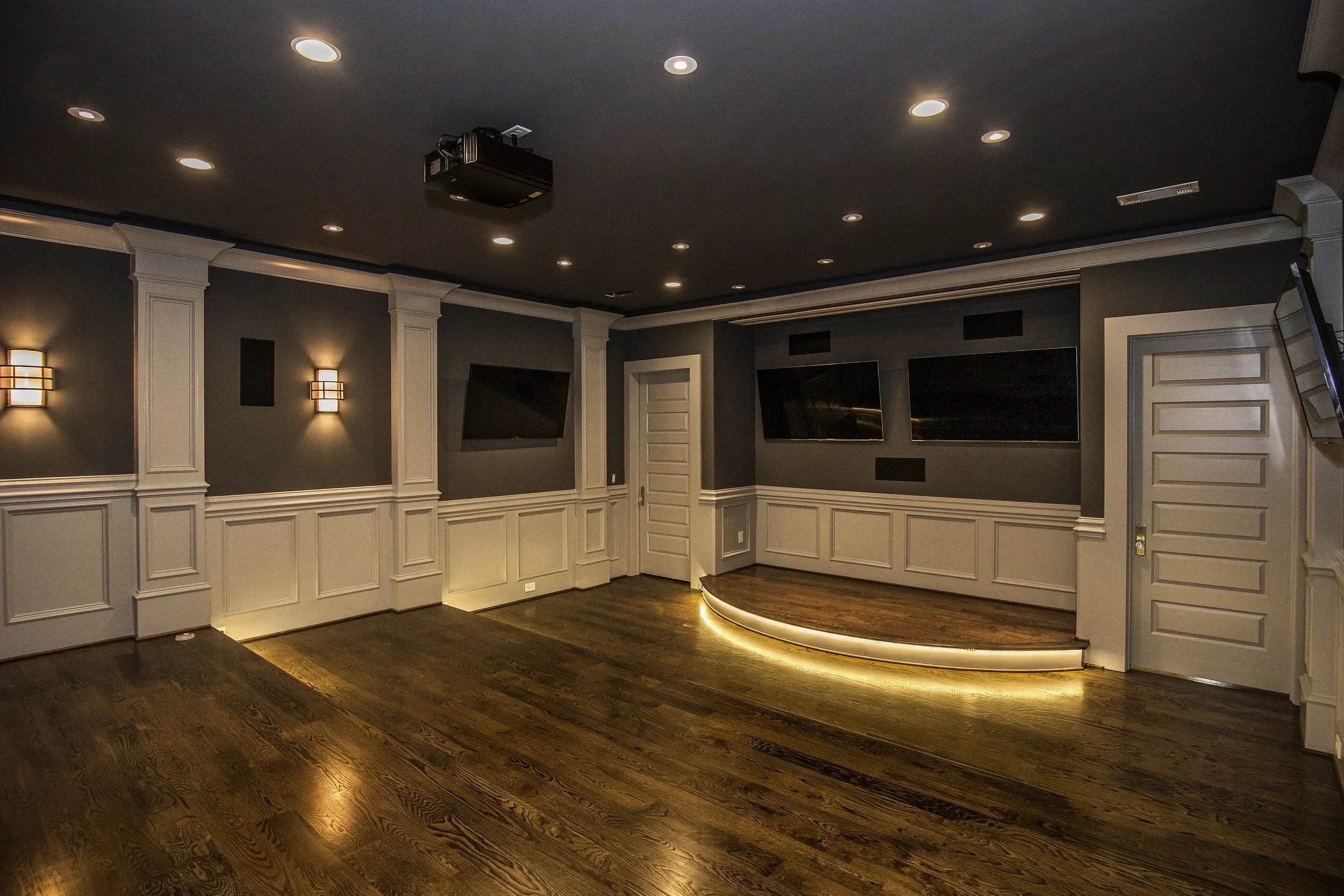 A finished basement room with dark hardwood flooring, white wainscoting, and a dark gray upper wall. It features two wall-mounted flat screen TVs, a curved accent wall with lighting underneath, and recessed ceiling lights. A projector is mounted on t