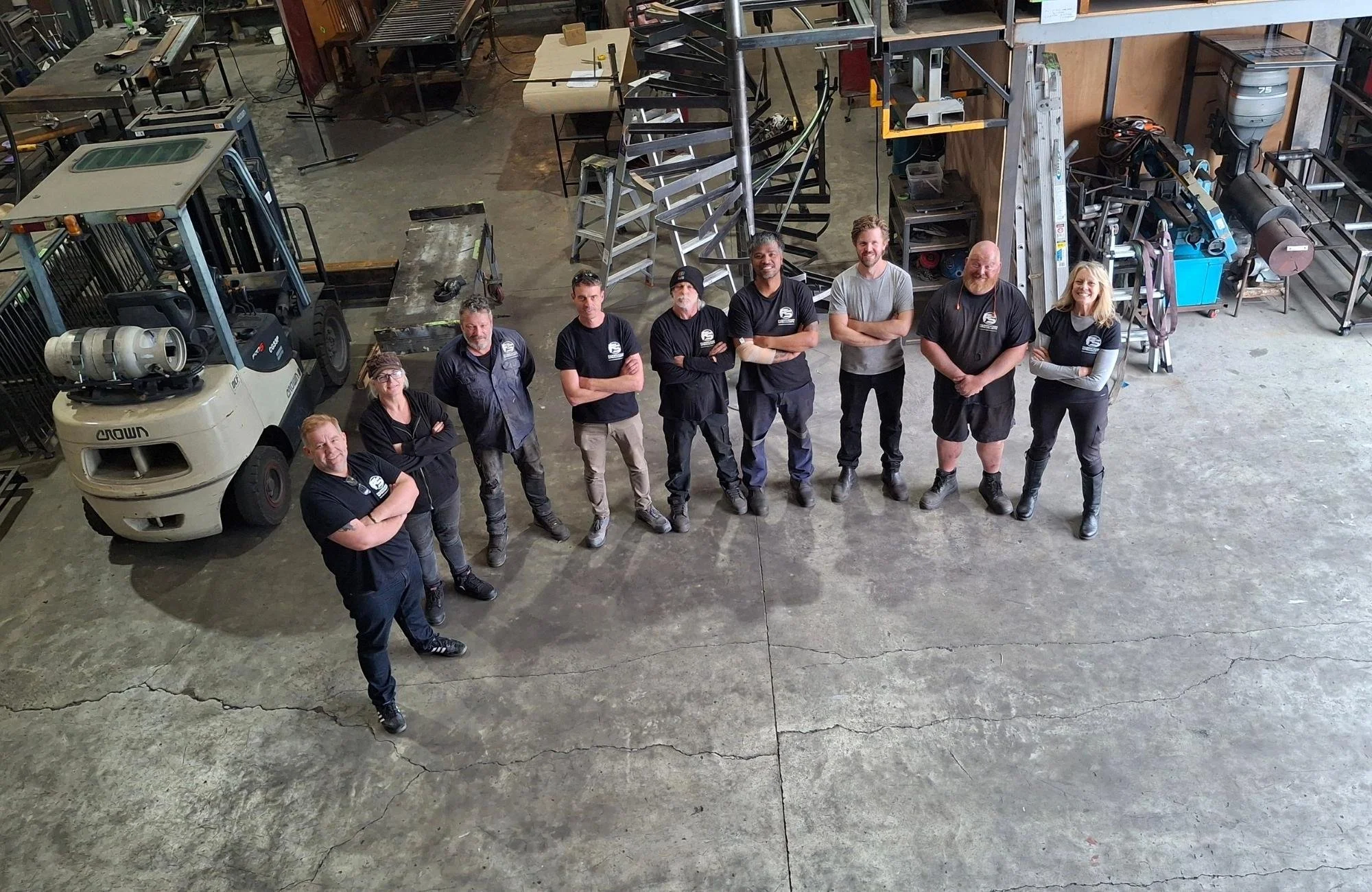 A group of eleven workers standing in a line inside a fabrication workshop. The background includes a forklift, metal ladders, workbenches, and various tools and equipment.
