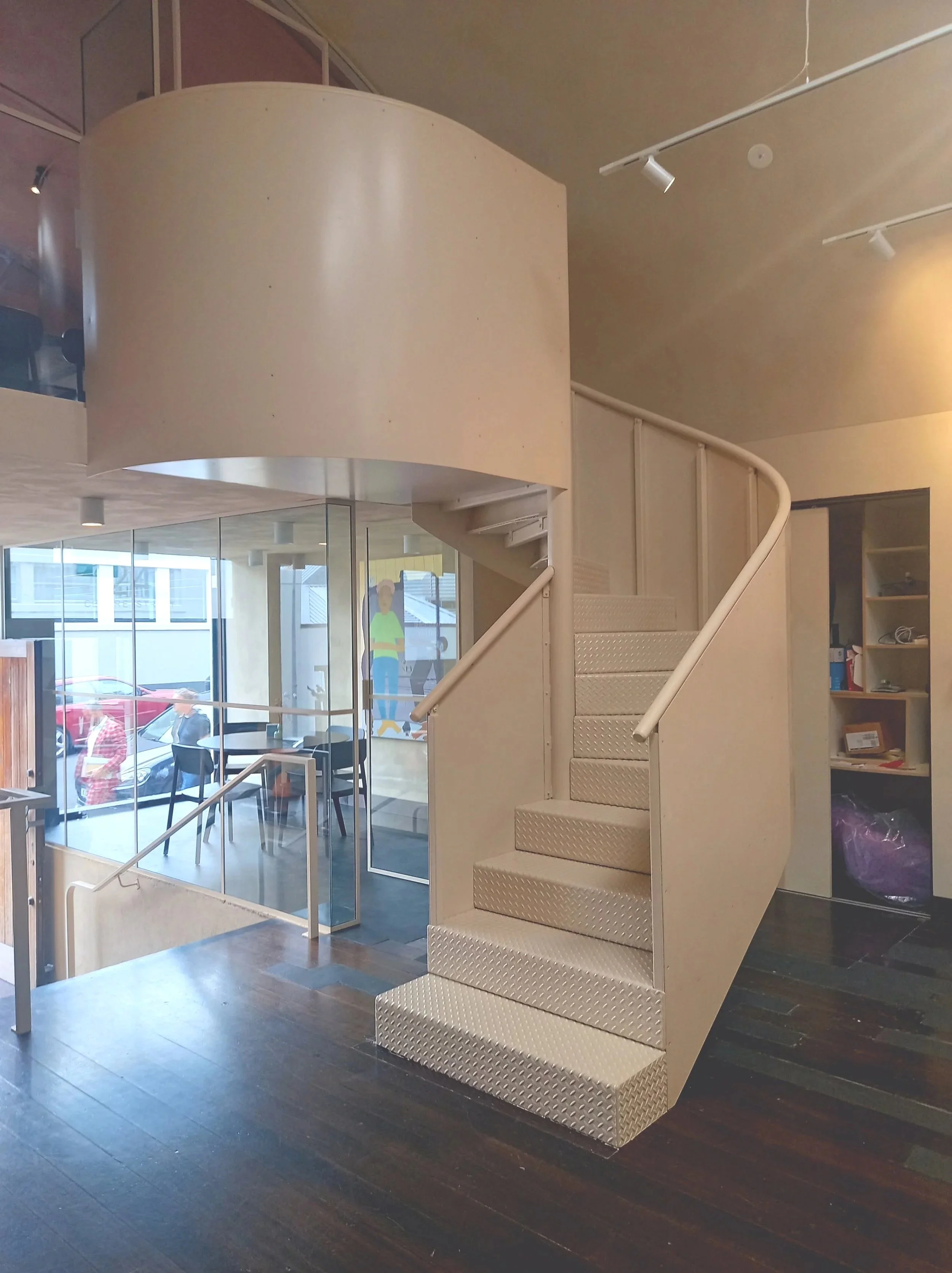 Interior of a modern building featuring a white curved steel staircase, glass walls, dark wooden floor, and a bookshelf.