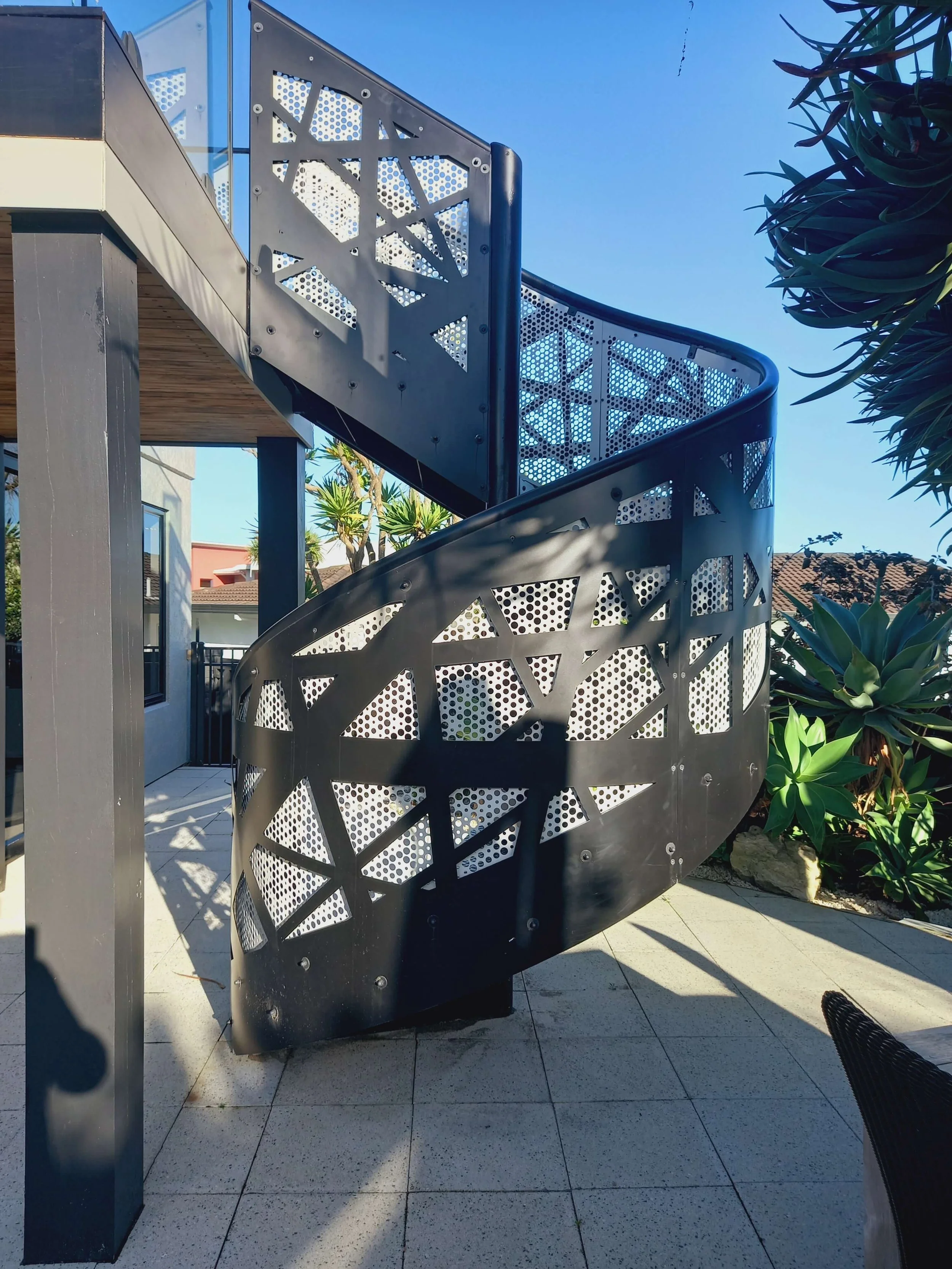 Outdoor metal spiral staircase with decorative cutout patterns, attached to a building with a wooden soffit, surrounded by plants and trees, under a clear blue sky.