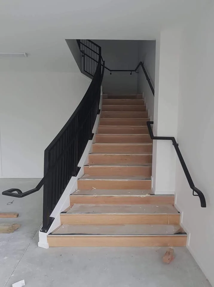 Wooden staircase under construction with black metal handrails in a white-walled building.