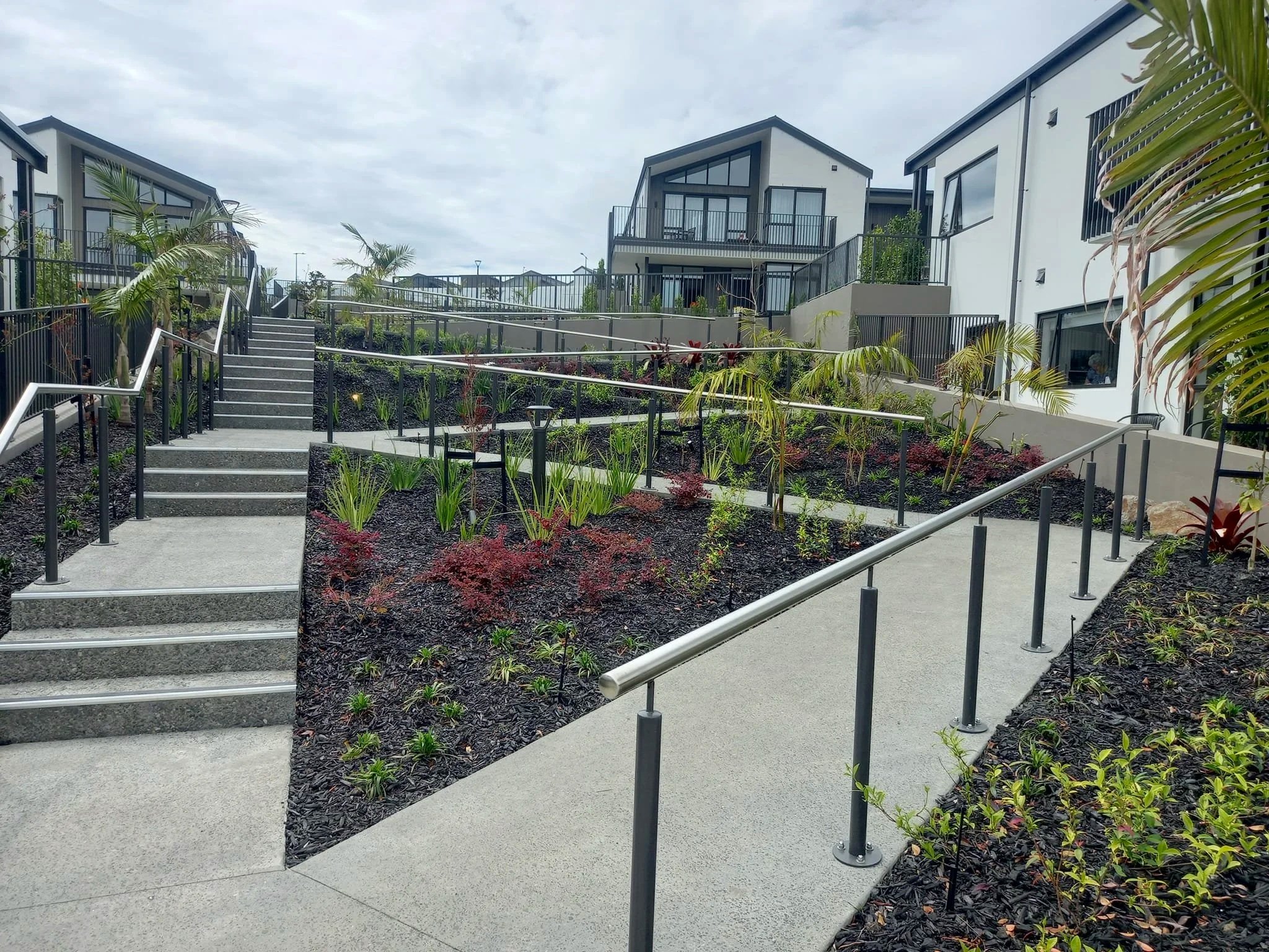 Outdoor stairs and ramp with metal railings in a modern residential area, landscaped with various plants and shrubs.
