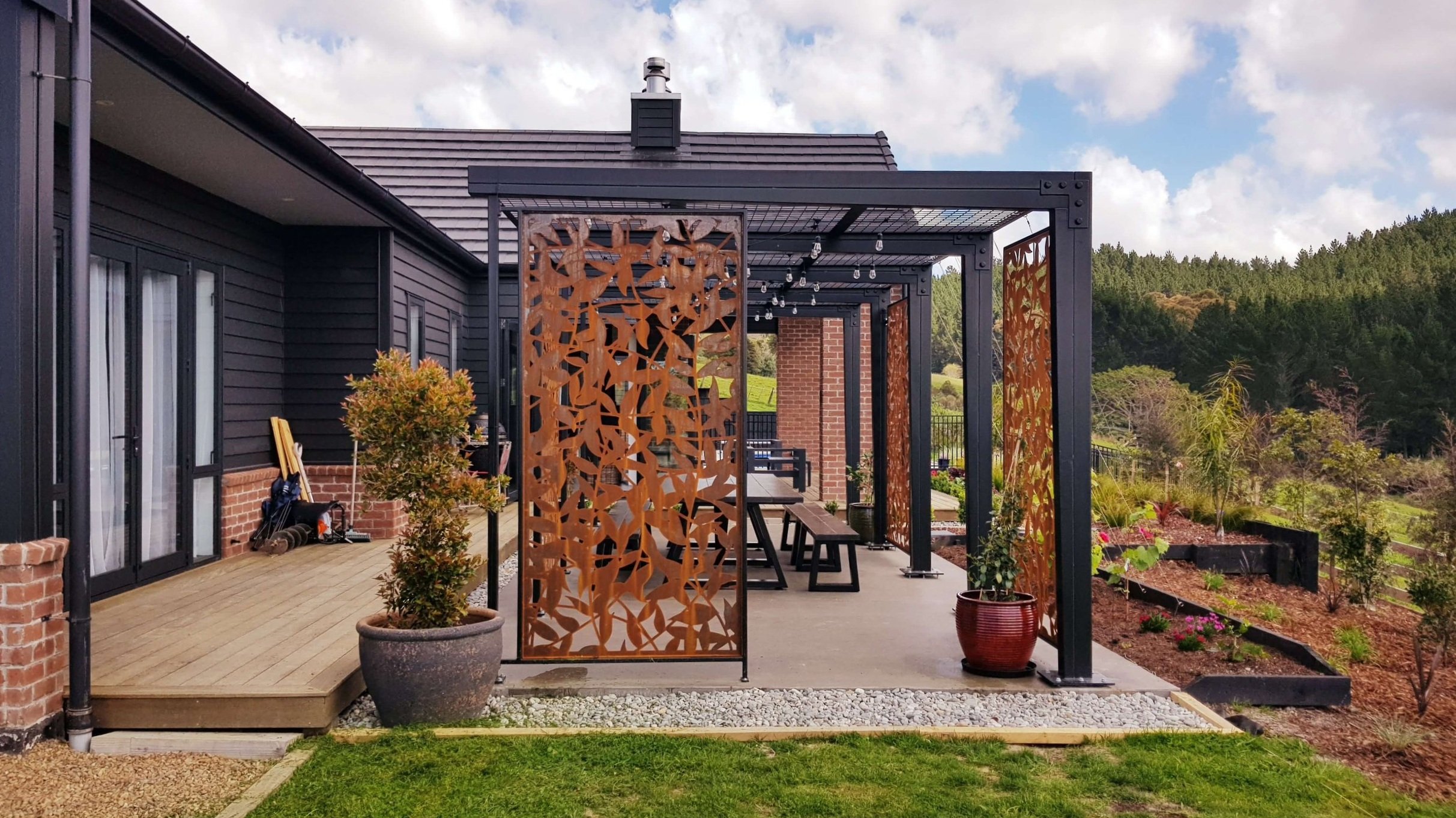 A modern outdoor patio area with decorative steel lattice panels, an outdoor dining table with benches, potted plants, and string lights, set against a background of green hills and overlooking a landscaped garden in Wainui.