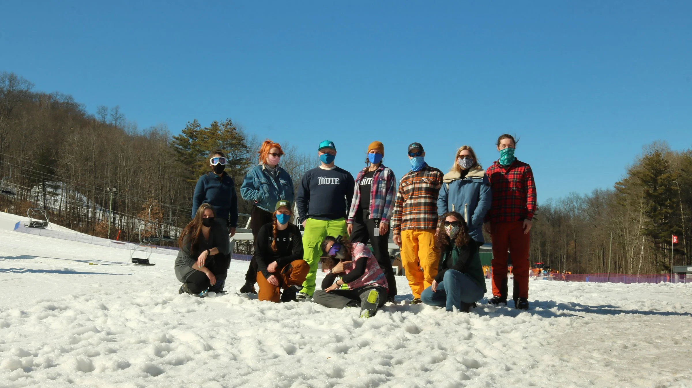A group of people wearing masks and winter clothing, standing and kneeling on snow in an outdoor ski resort area, with ski lifts and trees in the background.