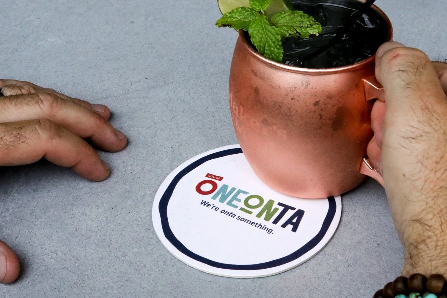 Drink in a copper mug being placed a coaster that has the Oneonta Logo on it
