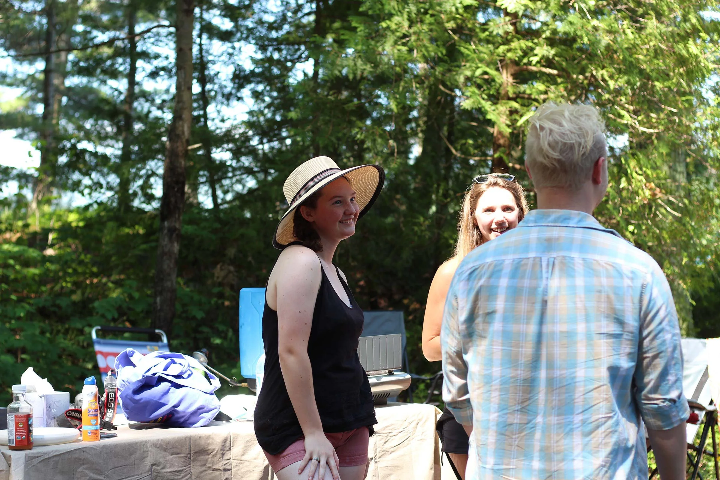 Group of people outdoors, two women smiling and talking, surrounded by trees, with a table that has food and drinks.