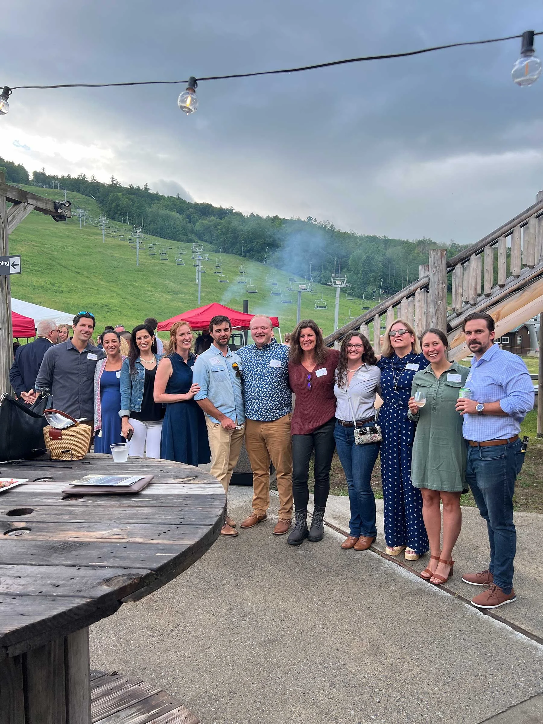 A group of people at an outdoor event with a mountain and ski lift in the background, under a cloudy sky.