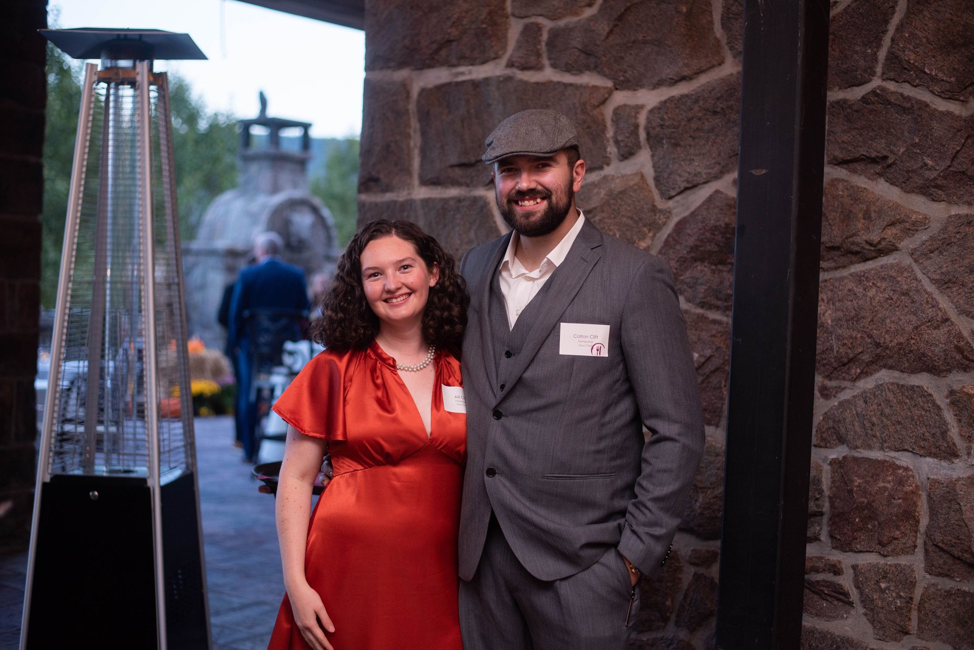 A man and woman standing together at an indoor event, smiling at the camera. The woman is wearing a shiny orange dress and pearl necklace, the man is in a grey suit with a white shirt and a flat cap. There is a stone wall behind them and a gas patio 
