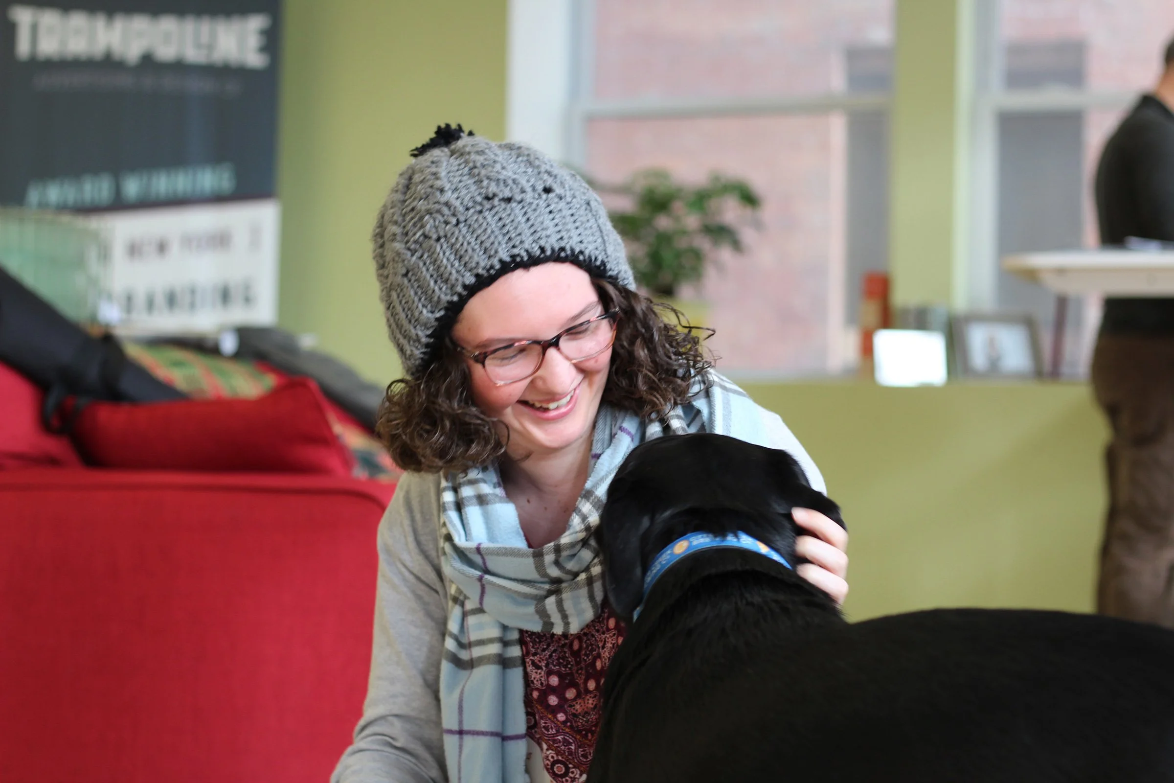 A woman with curly hair, glasses, and a knit hat smiling while petting a black dog wearing a blue collar in a room with green walls and large windows.