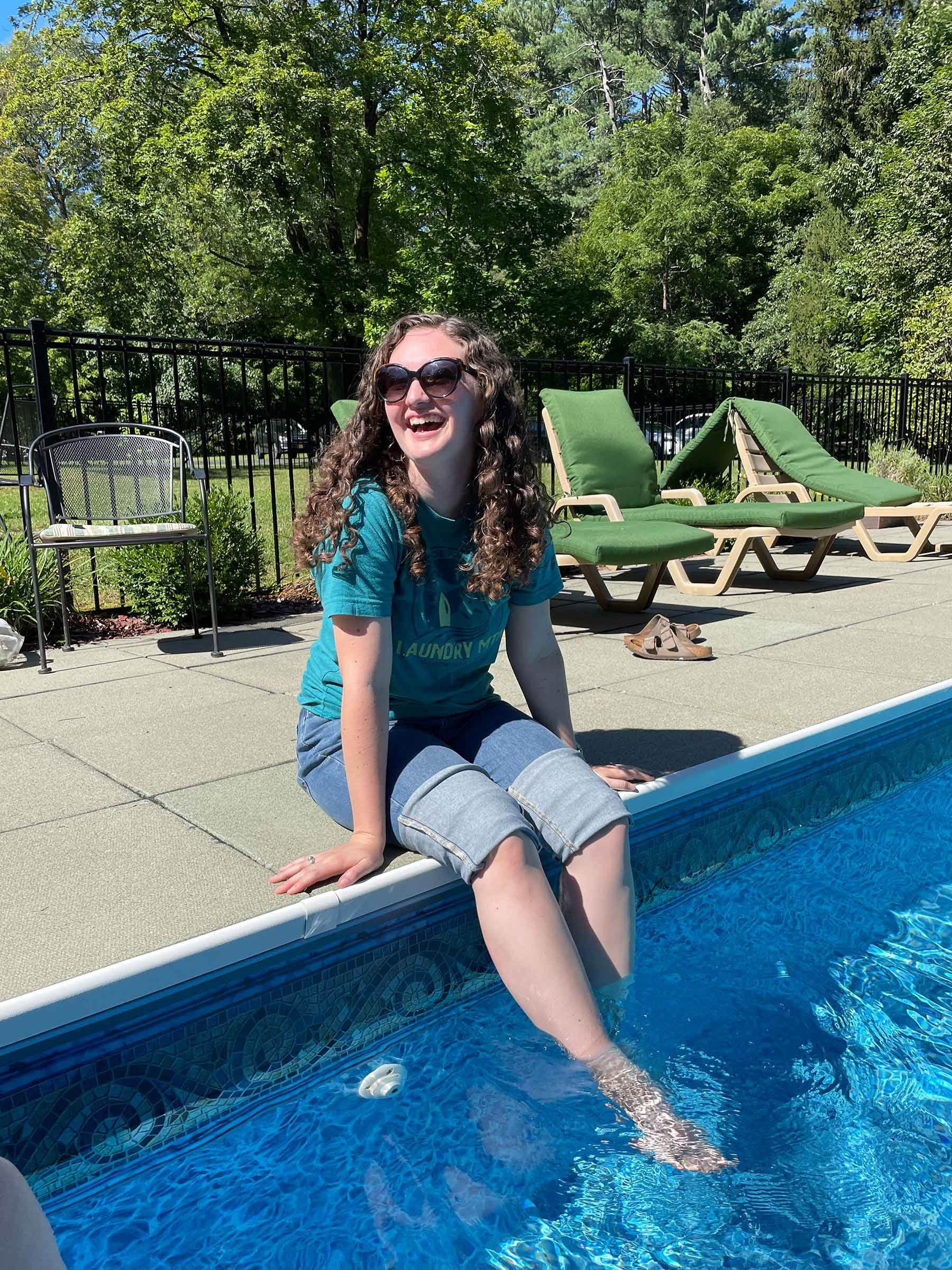 A woman with curly hair and sunglasses sitting at the edge of a swimming pool, smiling and enjoying a sunny day.