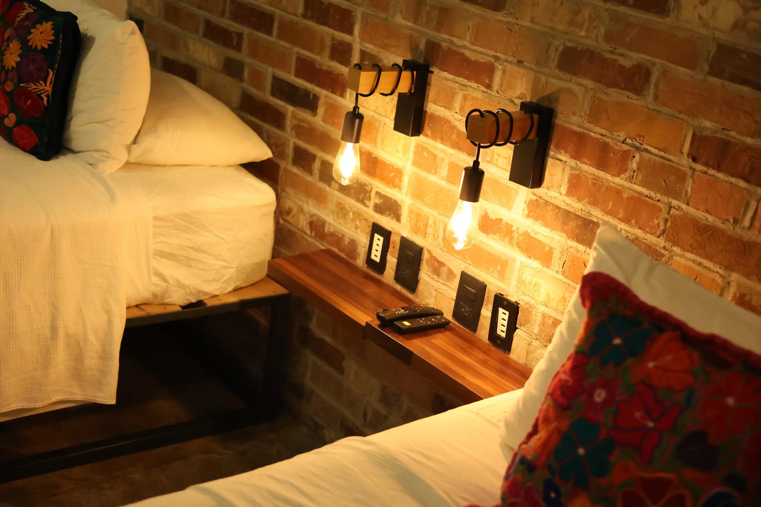 Hotel room with a brick wall, king-sized bed with white pillows and gray blanket, wall-mounted air conditioner, flat-screen TV displaying the Netflix logo, concrete ceiling, and plush beige carpet.