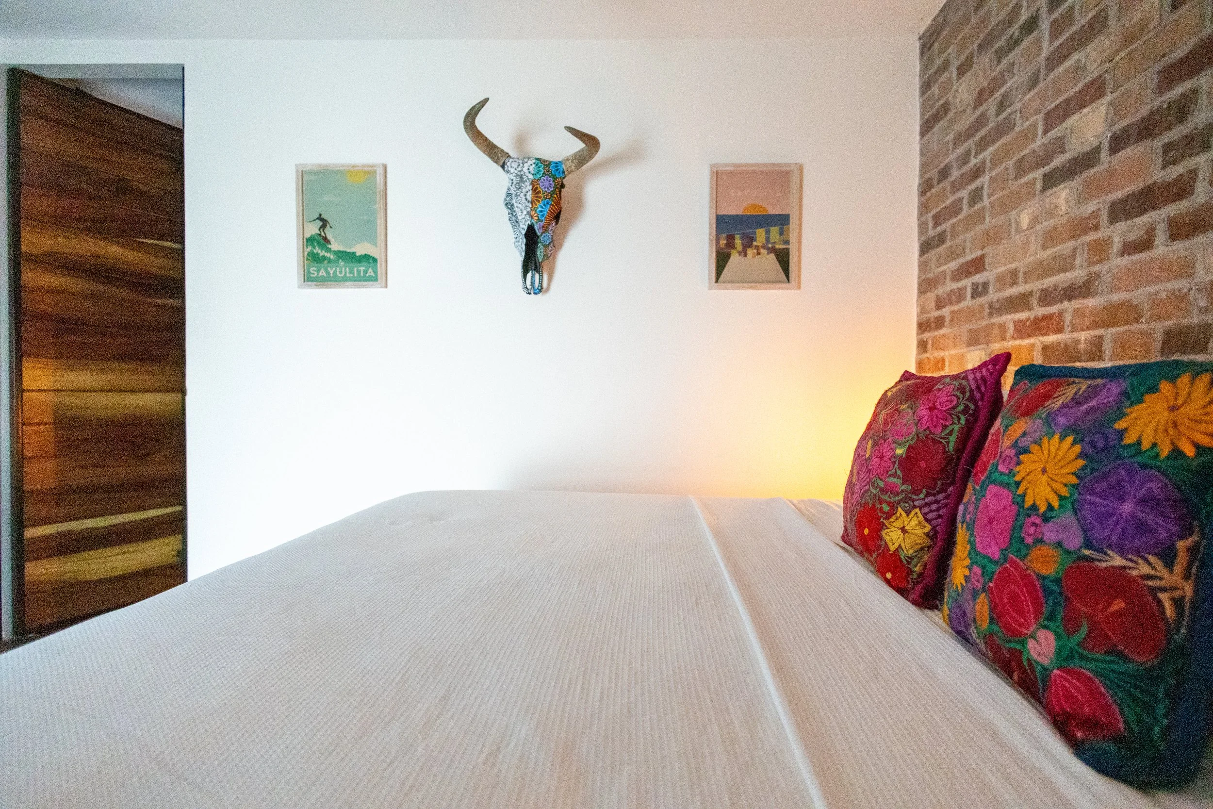 A cozy hotel bed with white sheets, pillows, a gray blanket, and warm wall-mounted lights on a brick wall in a rustic-style room.
