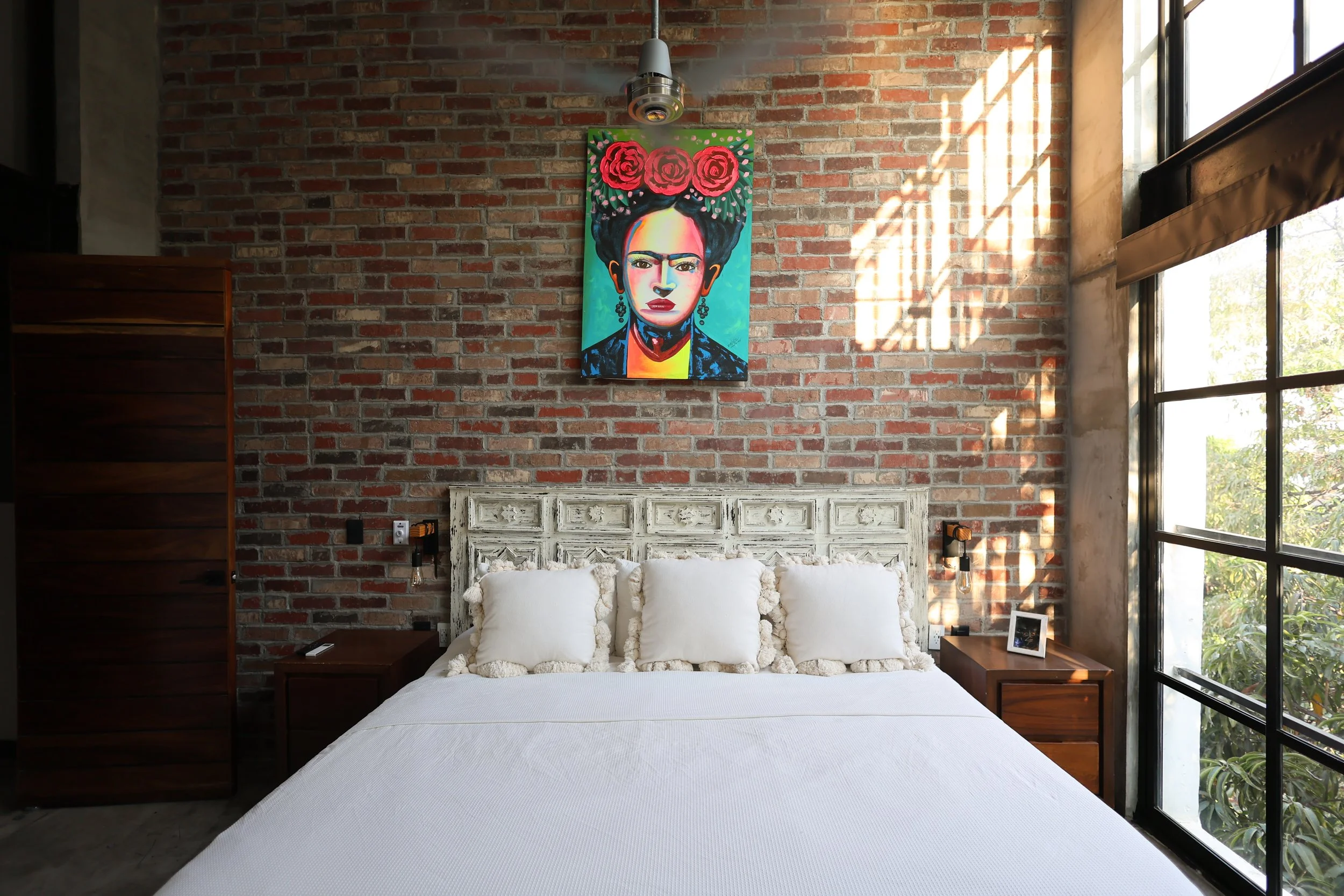 A modern bedroom with a large bed, dark gray bedding, ornate white headboard, brick wall behind the bed, and large floor-to-ceiling windows showing green trees outside.