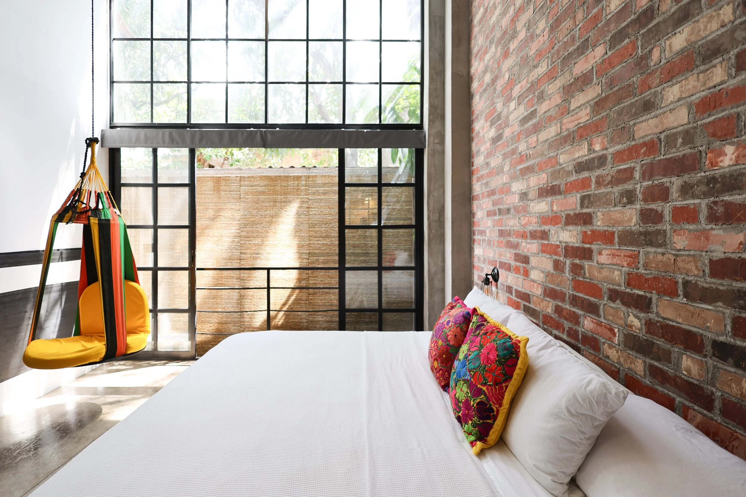 Modern bedroom with an exposed brick wall, large window, and colorful artwork above the bed. The room features a hanging chair with bright stripes, a white bed with gray bedding, wall-mounted lights, and an air conditioning unit.