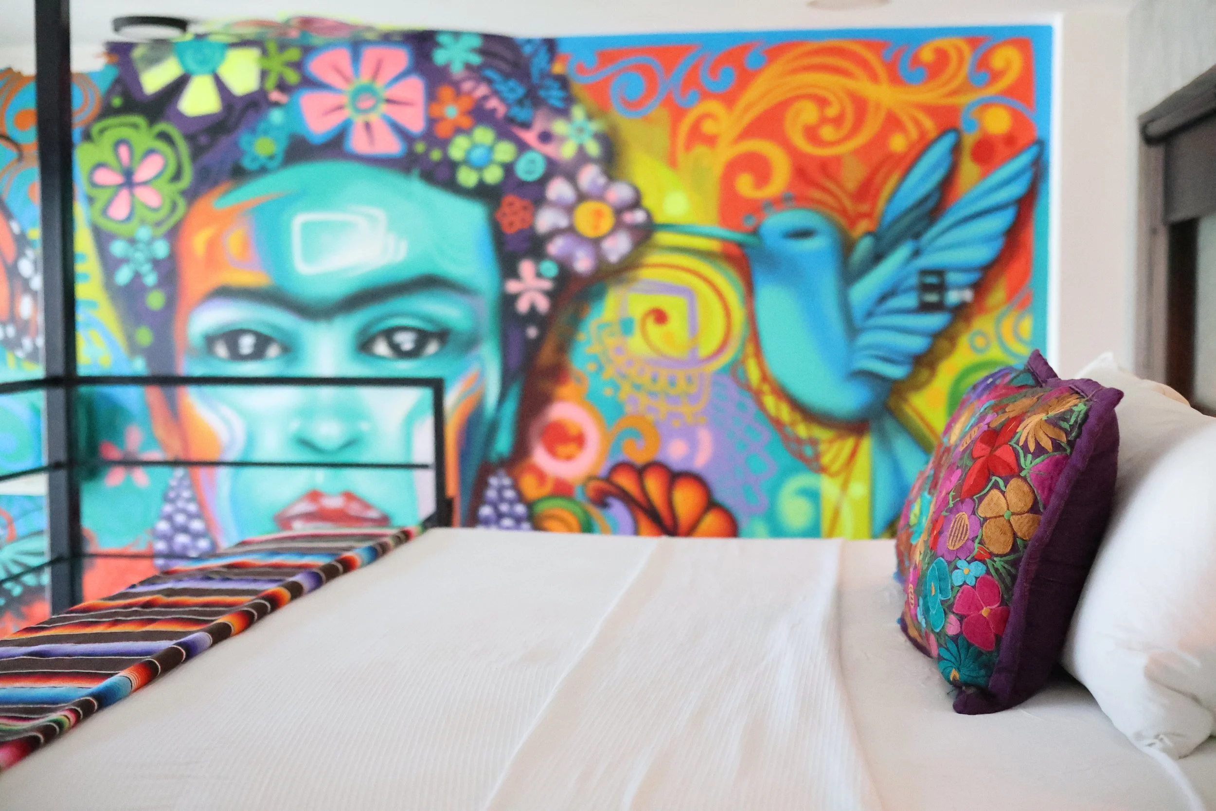 Modern bedroom with a white bed, gray blanket, brick wall, and colorful mural of a woman's face with flowers and butterflies.