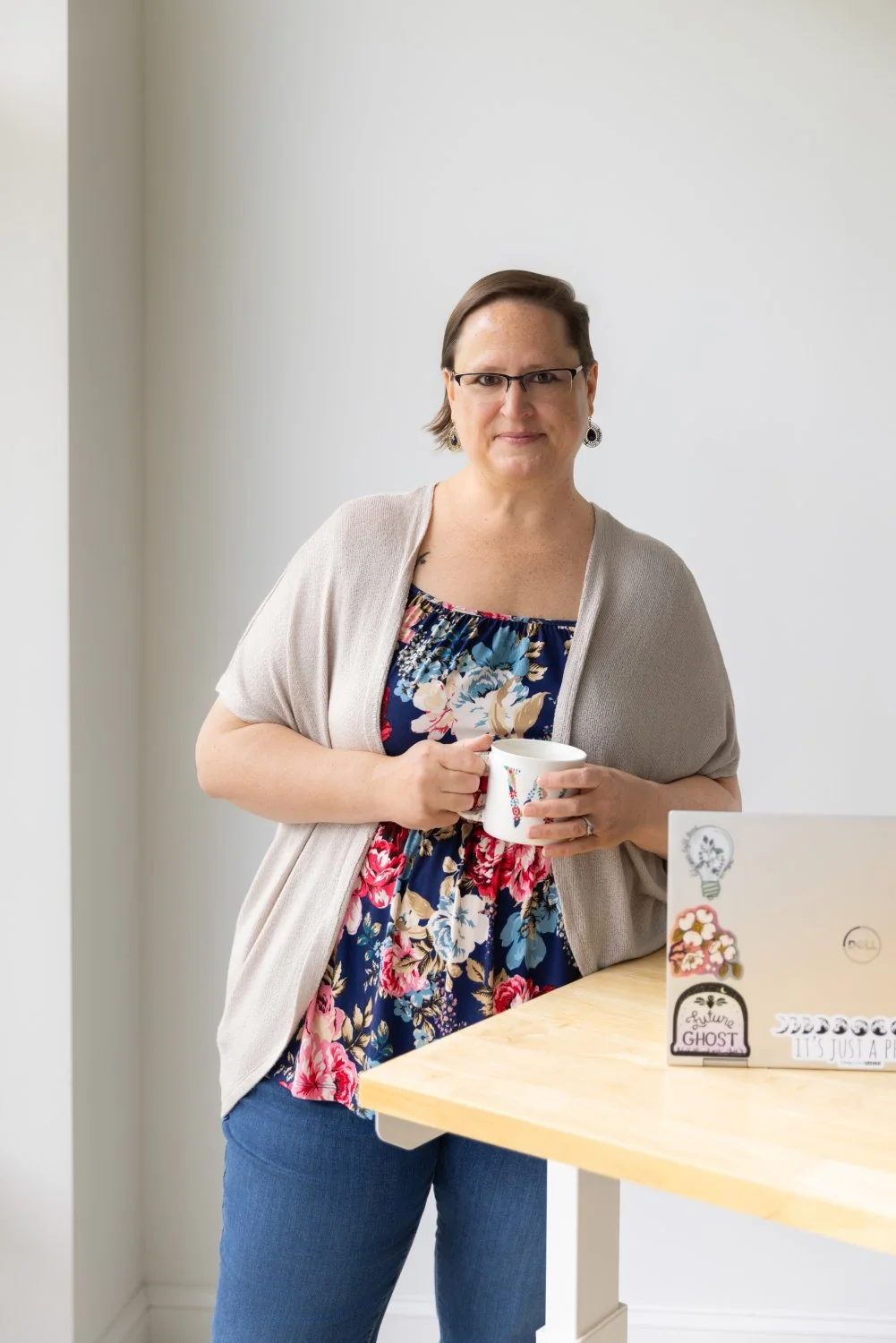 A woman with short brown hair, glasses, and earrings, holding a white mug, standing in front of a white wall and a light-colored table with a laptop decorated with stickers.