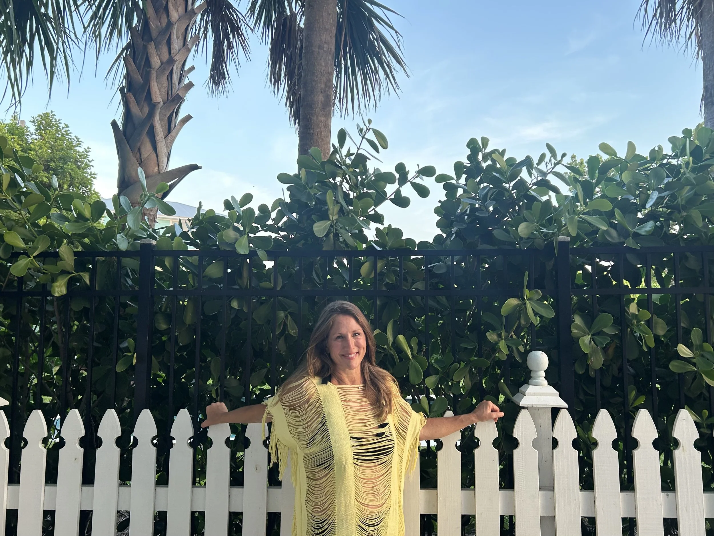Woman with long hair wearing a yellow fringed top, standing outdoors in front of a white picket fence, with green bushes, trees, and a blue sky with some clouds.