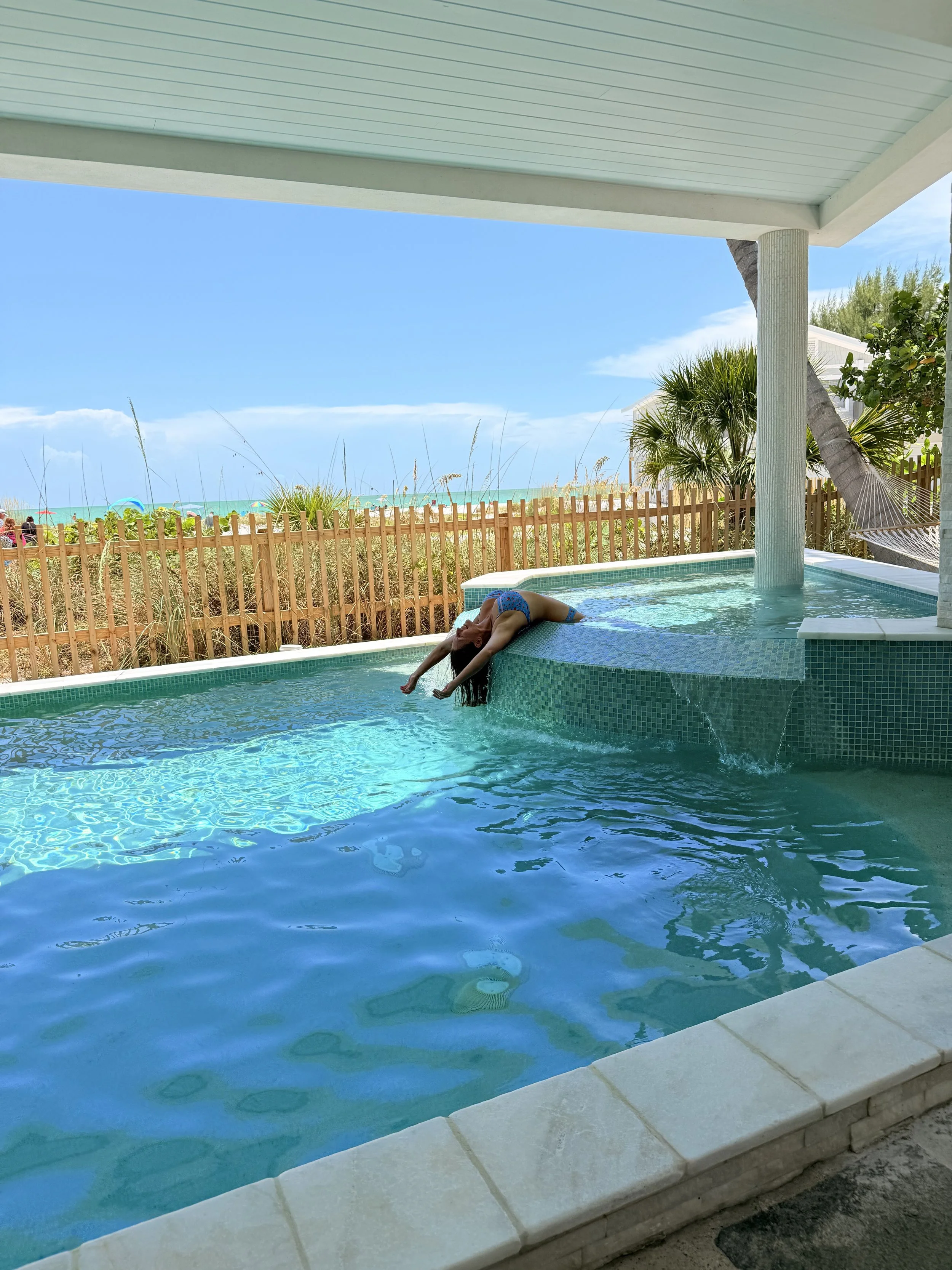 A woman is leaning over the edge of an outdoor pool, touching the water, with a beach and blue sky in the background.