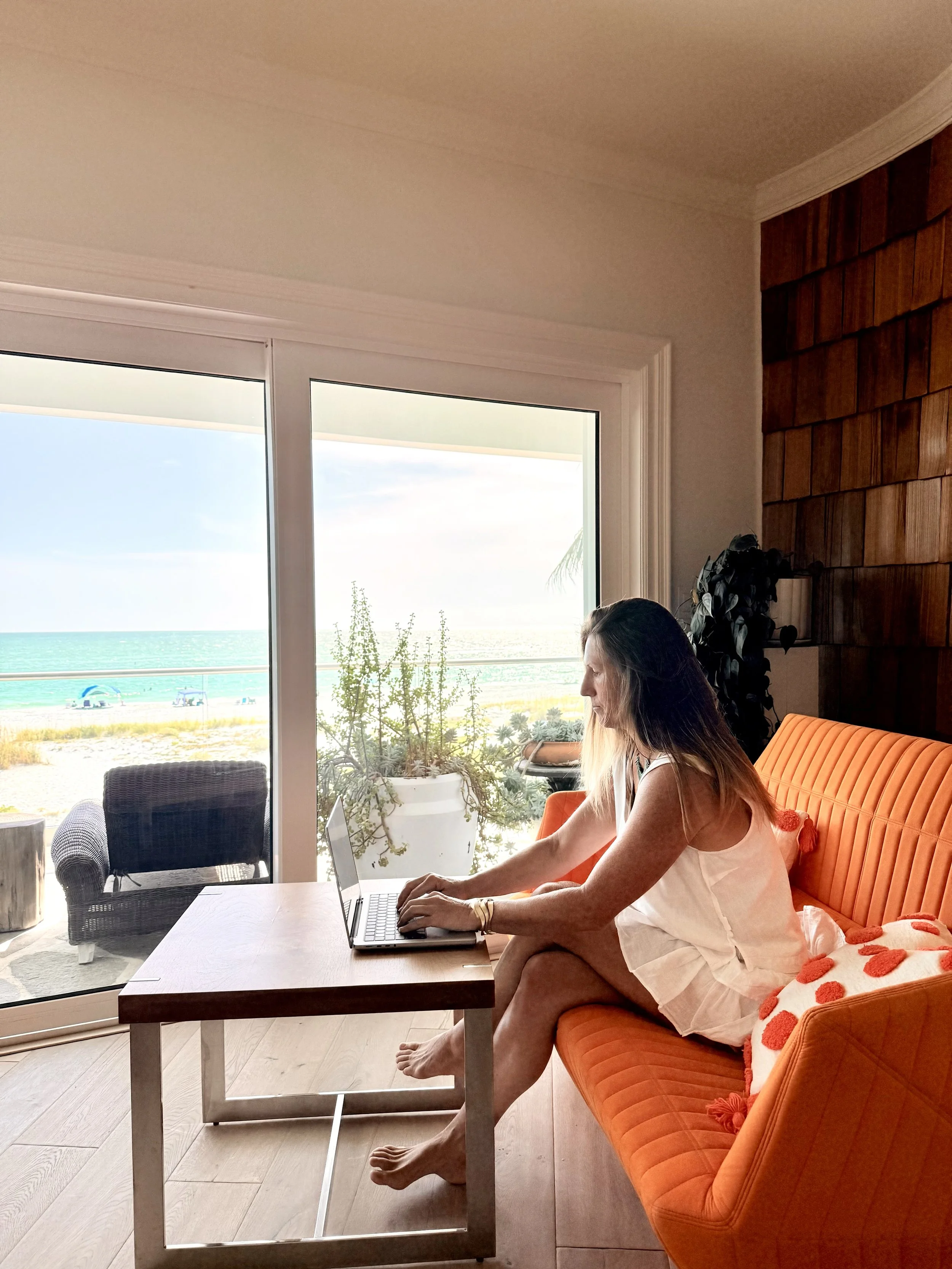 A woman with long hair and a white dress is sitting on an orange sofa, working on a laptop placed on a wooden table near a large window showing a beach view.