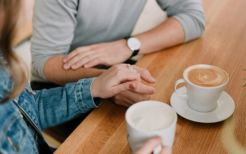 wei Menschen halten sich die Hände mit Kaffee-Tassen sitzend an einem Tisch