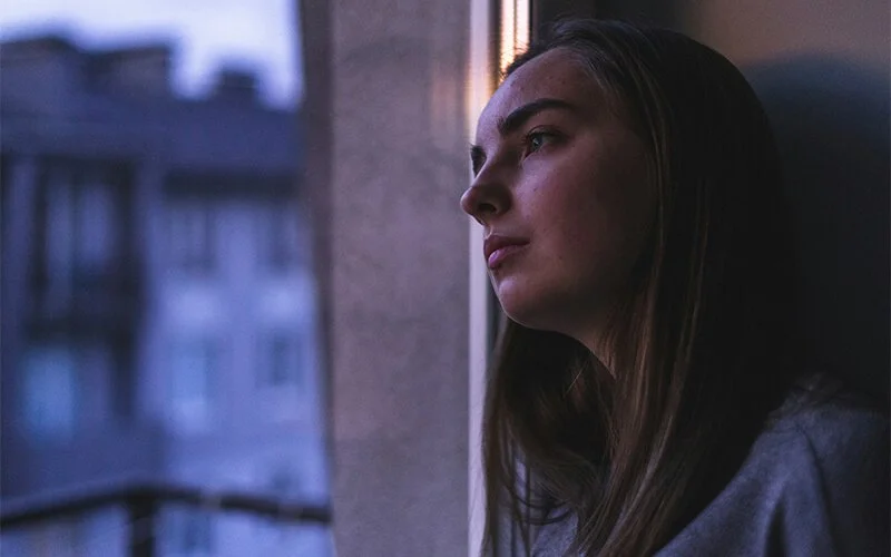 Eine weibliche Person lehnt nachdenklich an einer Fensterscheibe. Dahinter umschafft eine Häuserfassade
