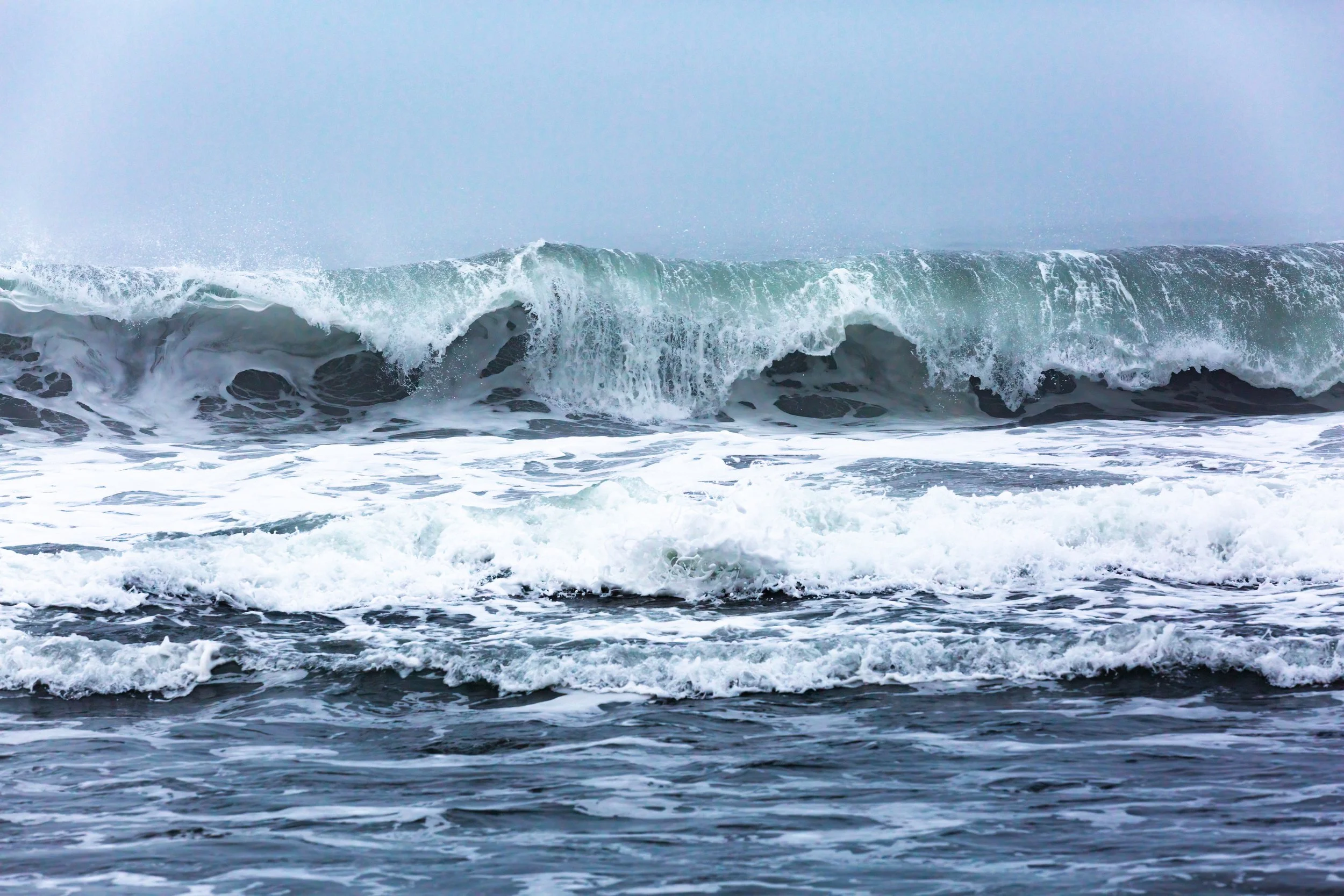 Large ocean wave is crashing with foam and spray in a cloudy sky.
