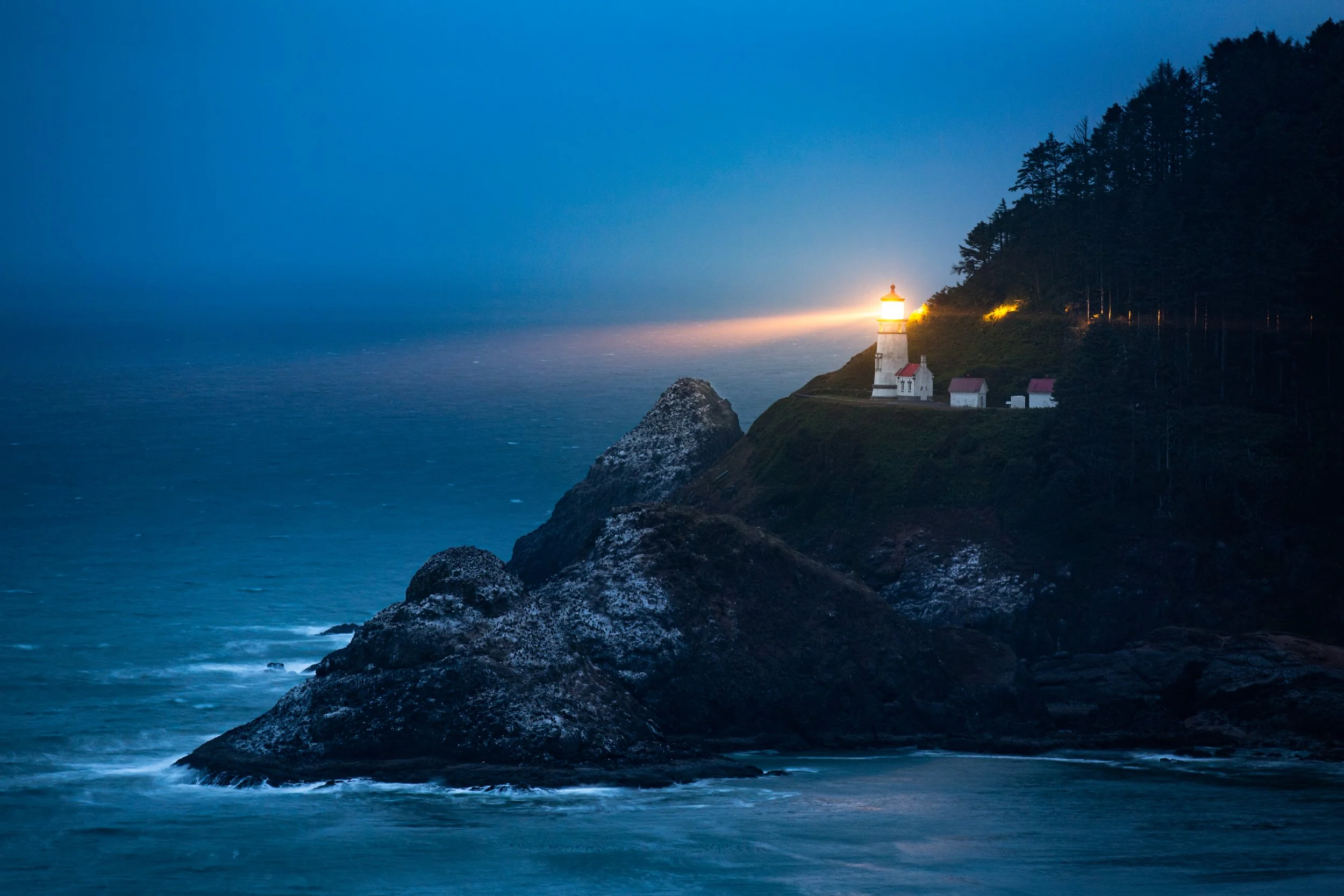 A lighthouse on a rocky cliff at dusk, with the light shining over the ocean, surrounded by trees.