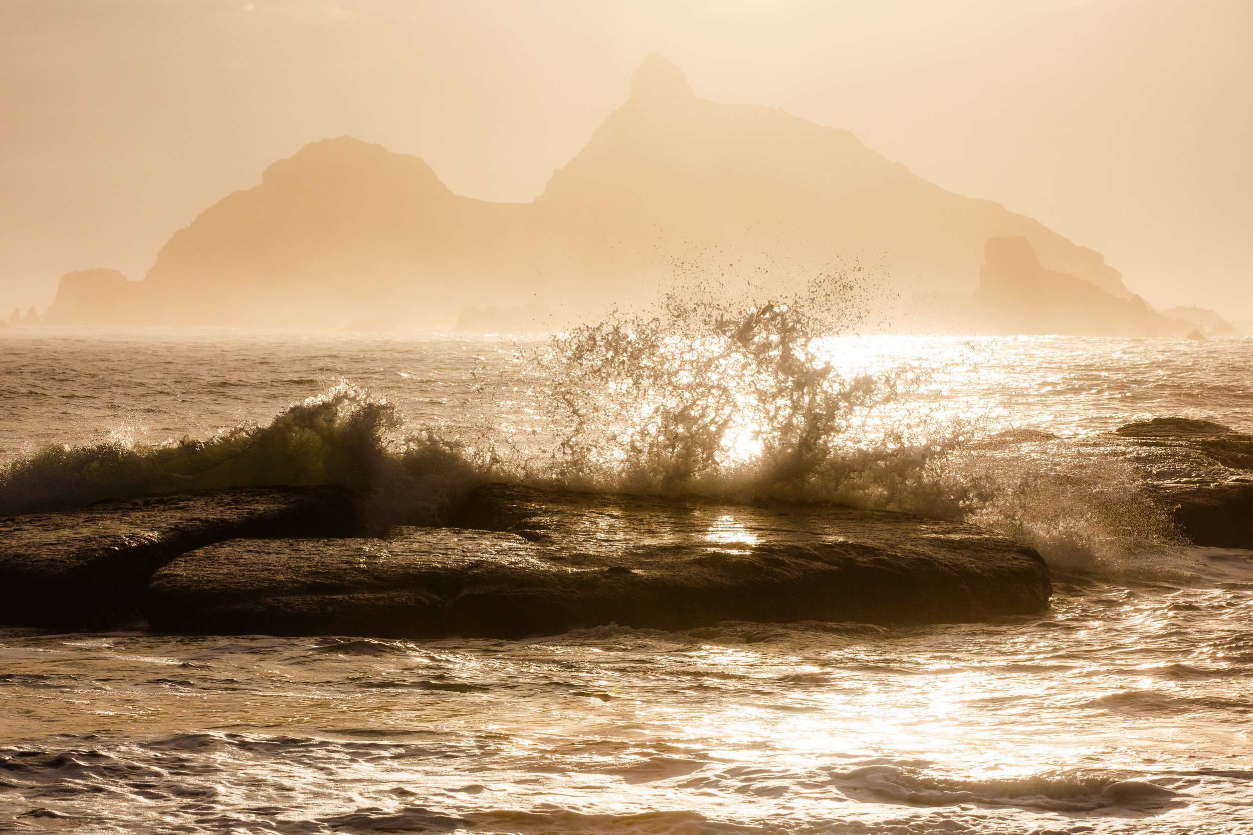 Sunset over the ocean with waves crashing on rocks and a distant mountain in the background.