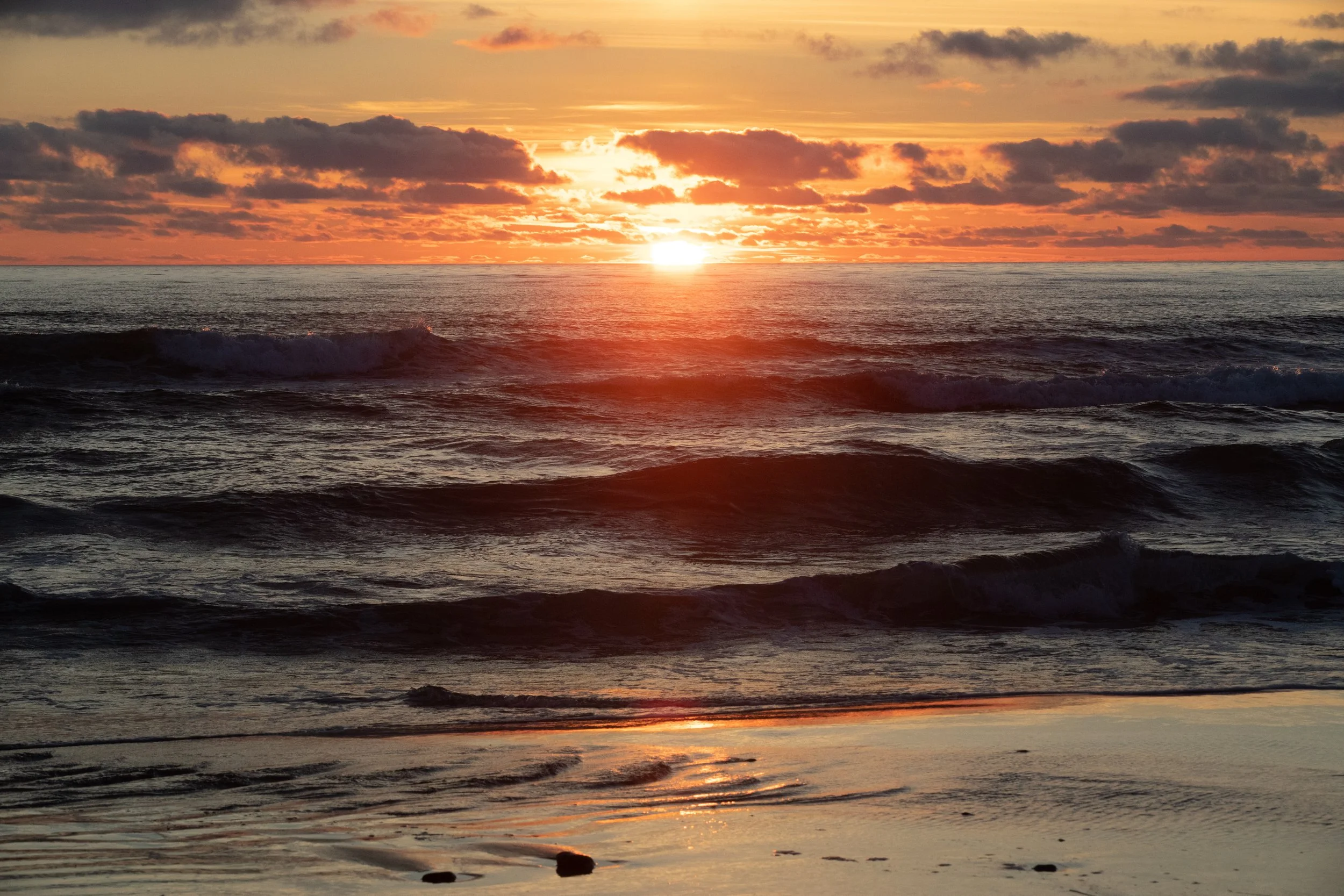 Sunset over the ocean with clouds, waves crashing on the shore, and reflections on the wet sand.