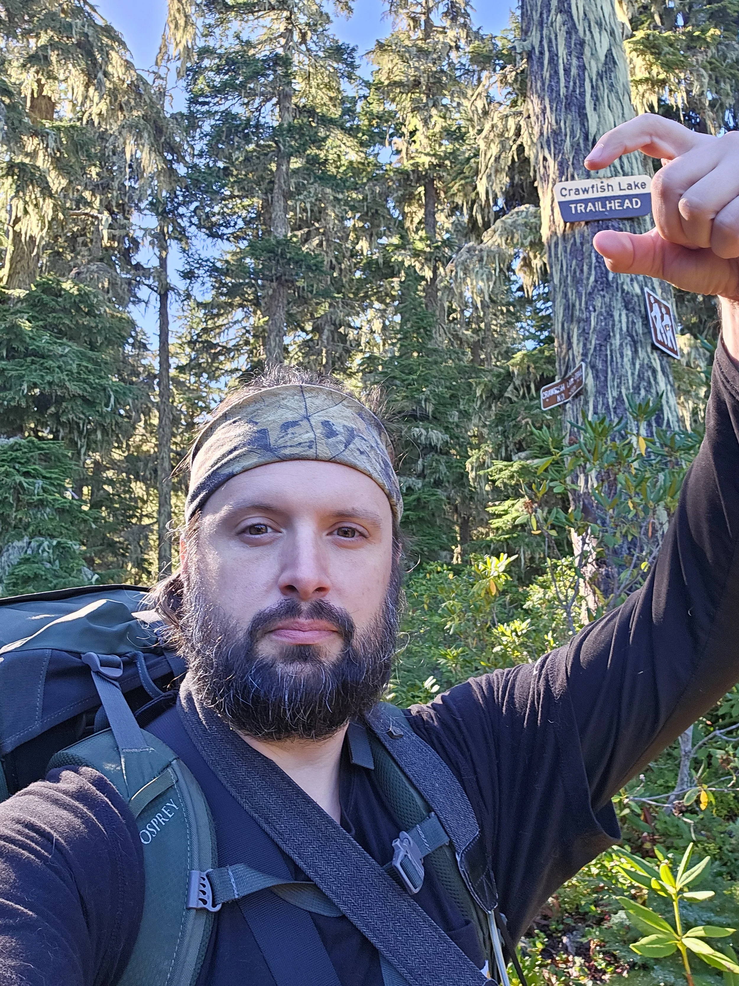 A man with a beard and a headband taking a selfie in a forested area, holding a trail sign that reads 'Crawfish Lake Trailhead.'