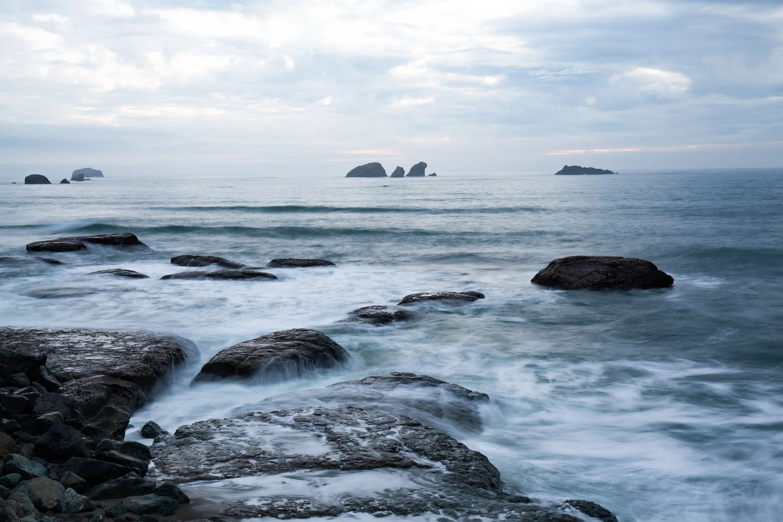 Long exposure photo of a rocky seashore with gentle waves and multiple distant rocks or small islands in the ocean under a cloudy sky.