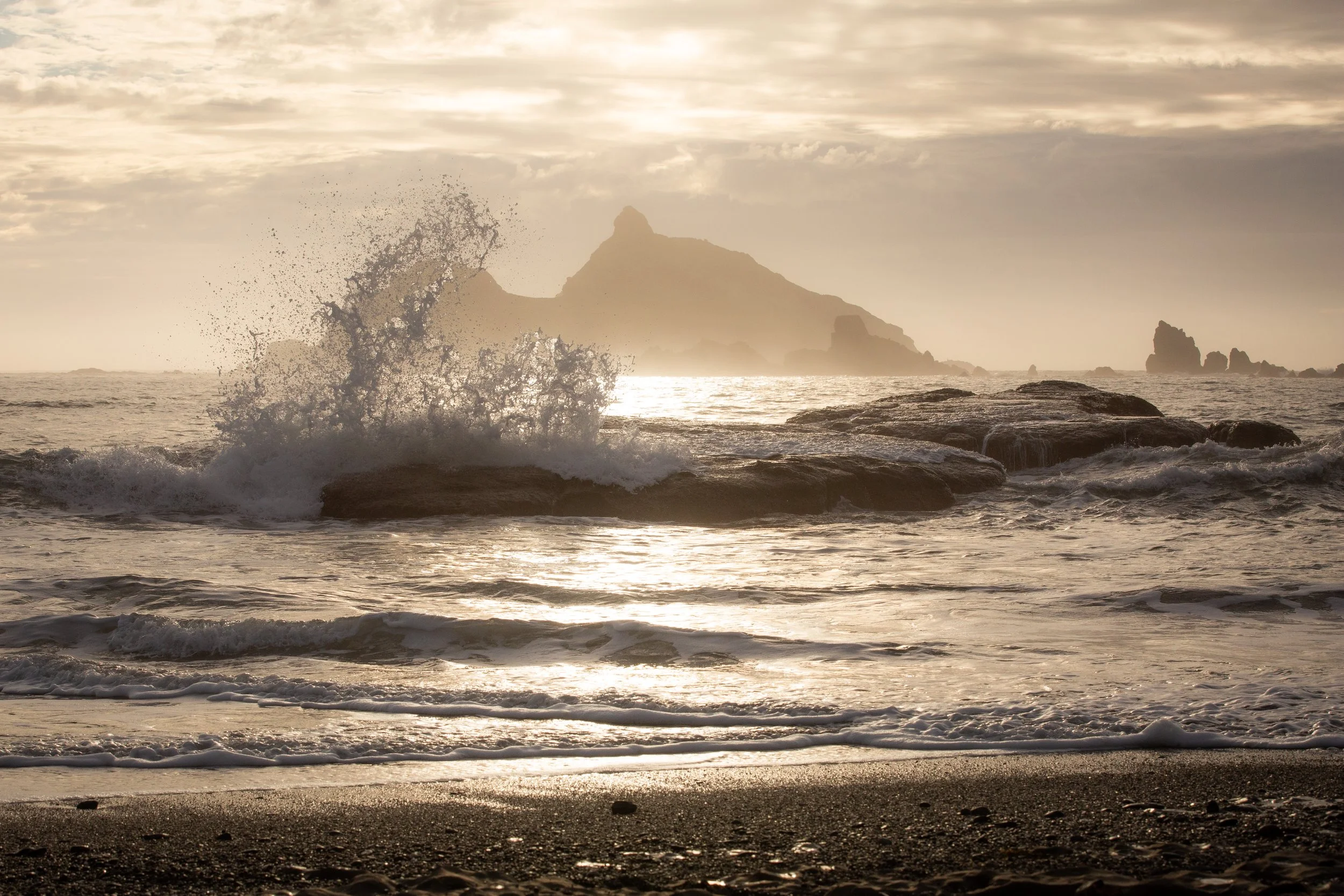 Sunset scene at the beach with waves crashing over rocks and a distant mountain in the background.